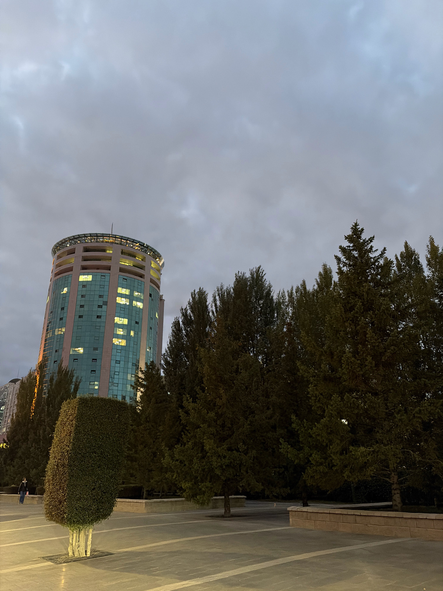 The image depicts a cylindrical, modern office building in Astana, Kazakhstan, under a cloudy sky. The building features a distinctive rounded top with a glass structure and is illuminated from within. Its facade is a combination of beige panels and teal-tinted glass windows, some of which are lit. In the foreground, a meticulously sculpted, rectangular hedge stands next to a paved plaza. A person, seen from behind and to the left, walks away from the viewer. Tall, dark green coniferous trees line the area between the building and the plaza, creating a dense visual barrier. The lighting suggests it is either dusk or a heavily overcast day.
