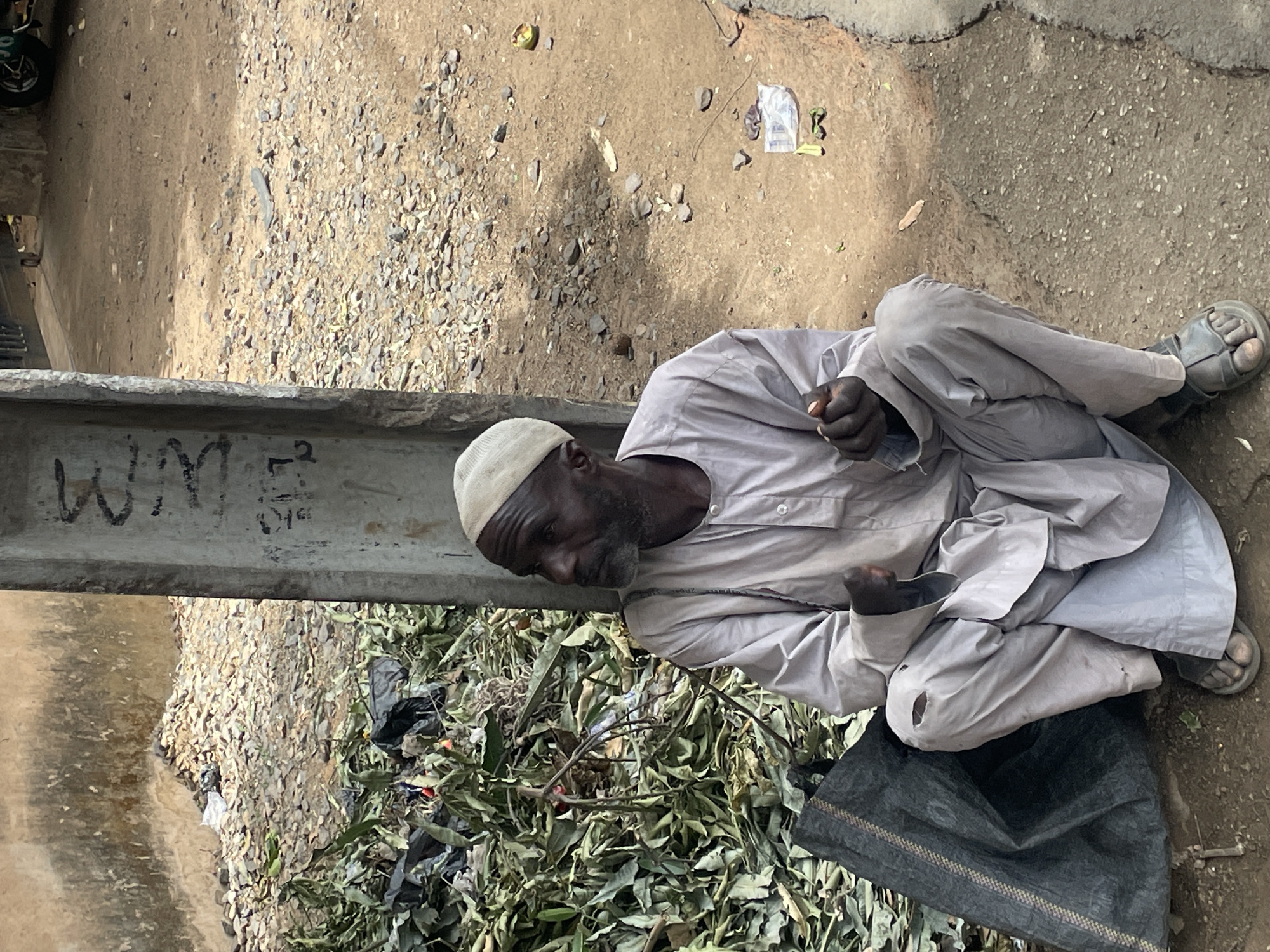 An outdoor photograph captures an elderly, dark-skinned man seated on a dusty, gravelly ground surface. He wears a light-colored traditional long tunic and a matching skullcap, with simple open-toed sandals on his feet. His hands are loosely clasped in front of him, and his gaze is directed downwards, suggesting a moment of quiet contemplation or weariness. A small tear is visible on the right knee area of his tunic.
The setting appears to be a street or pathway, characterized by a mix of dirt, small rocks, and patches of paved ground. Bright daylight casts strong shadows, indicating sunny weather. Behind the man, a sizable pile of dried green leaves, branches, and dark plastic debris is present. A large concrete beam or barrier cuts across the lower left of the frame. On this beam, the white letters "WM" are clearly visible, along with some fainter markings like "5^2". Various pieces of litter, including plastic bags and organic remnants, are scattered across the ground. The scene conveys a candid moment of everyday life in a simple, outdoor environment.