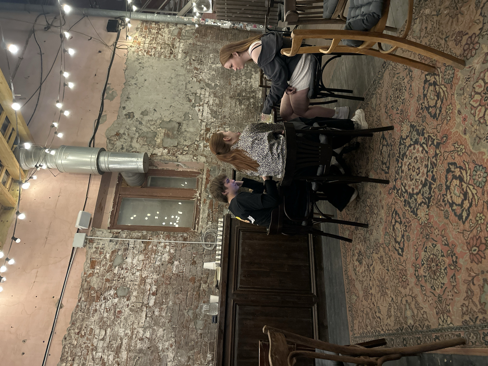 People sit and stand at a table indoors. The environment includes a brick wall, string lights, a silver metal pipe, a wooden door, and a patterned carpet. The location is not identified.