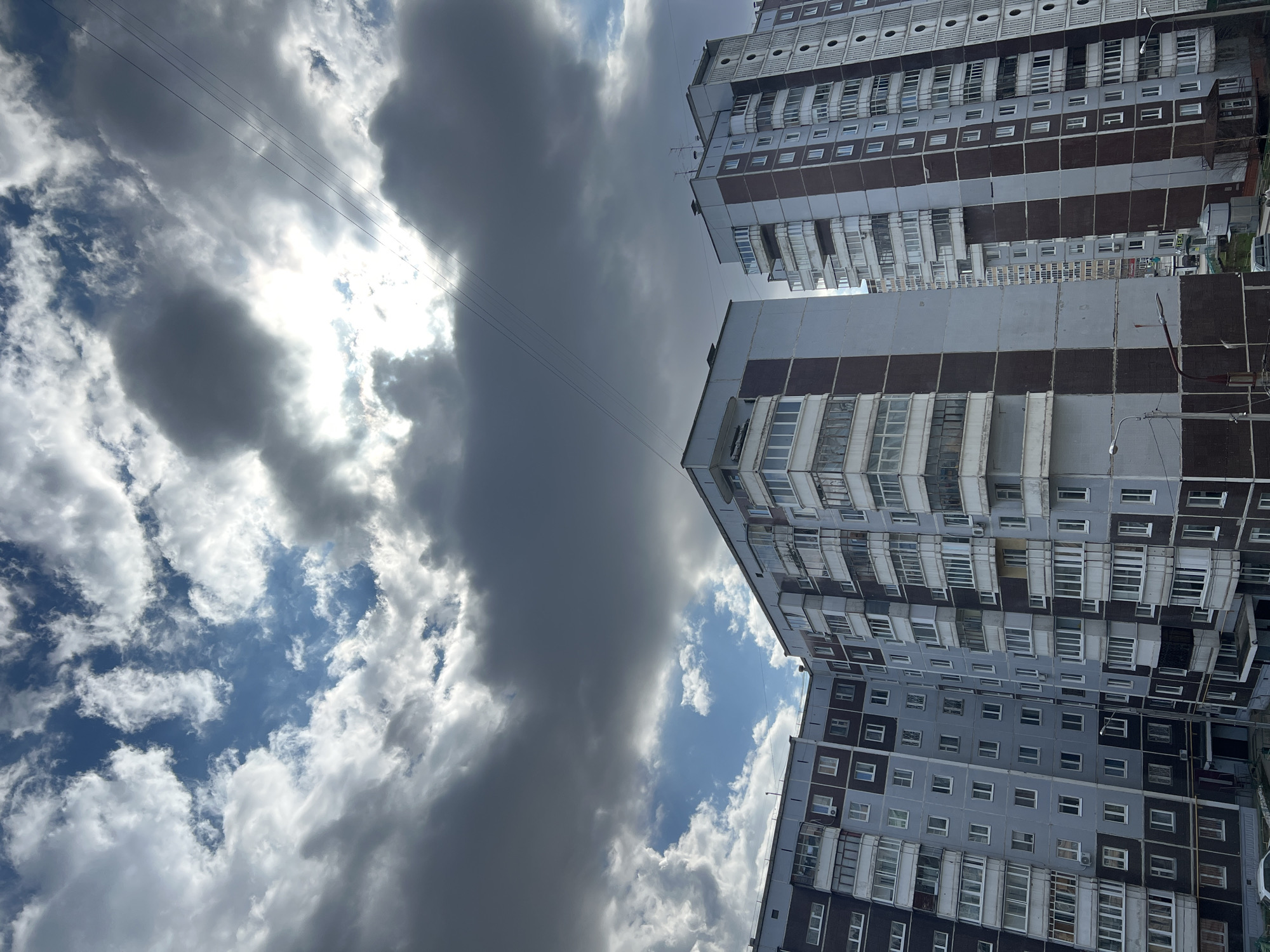 The image captures an upward-looking urban scene from Perm, Russia, featuring two tall residential buildings on the right and a dynamic, cloud-filled sky on the left. The multi-story apartment blocks display a modular architectural style with numerous windows and a mix of enclosed and open balconies. Their facades are characterized by alternating sections of light grey and dark reddish-brown panels. On the left side, the sky is dominated by large, textured clouds. Bright sunlight intensely breaks through a gap in the clouds in the upper-left portion of the frame, creating strong contrast with the darker, heavier cloud masses elsewhere. Several thin power lines stretch across the mid-ground of the sky. The lighting suggests a midday setting with partly cloudy weather, showcasing a common urban environment under a dramatic sky. No people or specific activities are visible, and there is no discernible text.