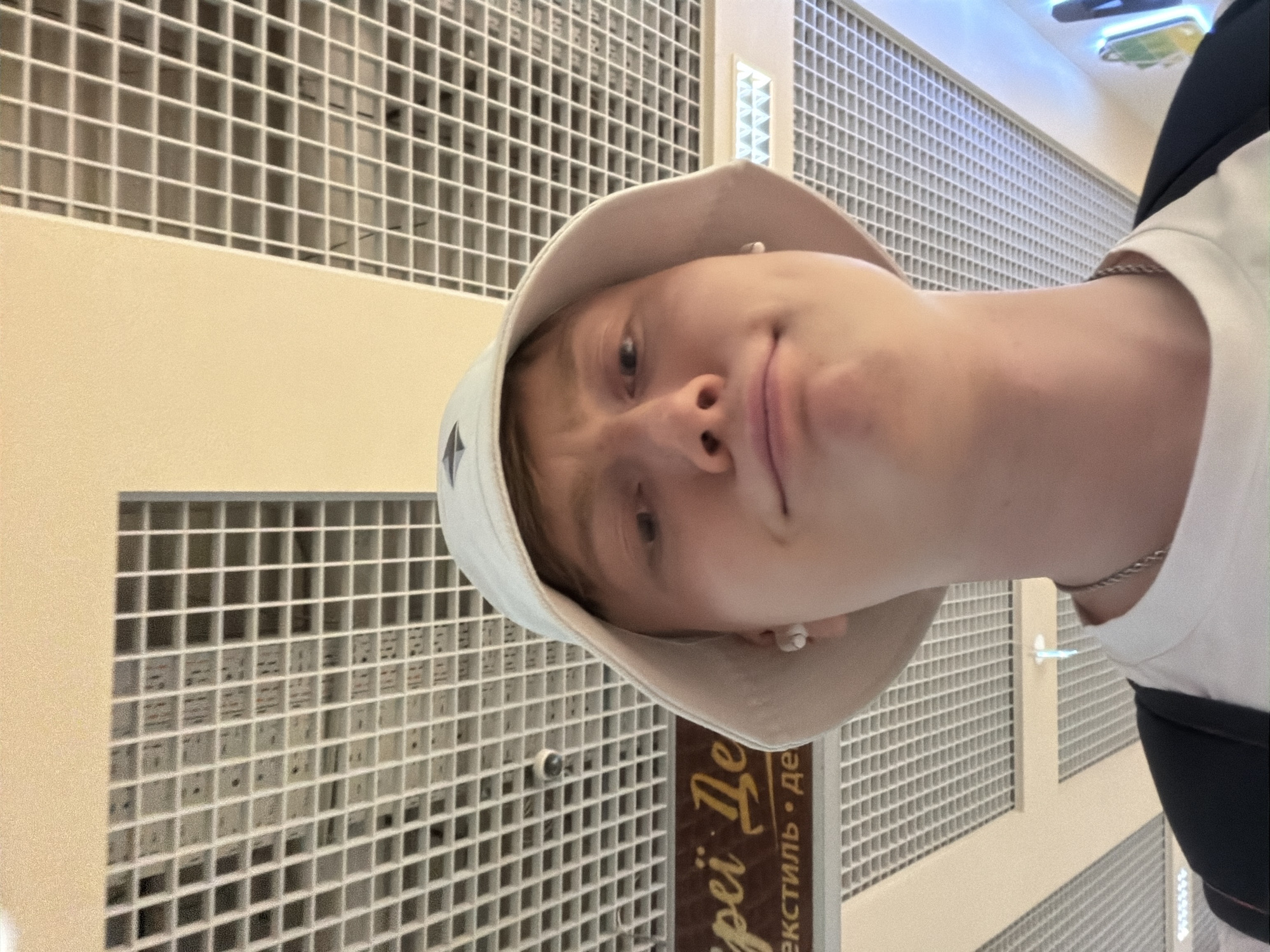 A young male with light skin, fair hair, and blue eyes is taking a low-angle selfie in an indoor location. He is wearing a light-colored bucket hat, a white t-shirt, a silver chain necklace, and a dark backpack. He has small white stud earrings in both ears. The background consists of a white drop ceiling with a grid pattern, characteristic of many shopping centers or public buildings. To the left of the subject, there is a brown sign with text in Cyrillic script. The text appears to be "arei Де" and "екстиль • Де". This signage, along with the architectural style of the ceiling, suggests the location is a shopping center, potentially in Kyiv, Ukraine, as the Cyrillic script indicates. The lighting is bright and evenly distributed, typical of an indoor commercial space.
