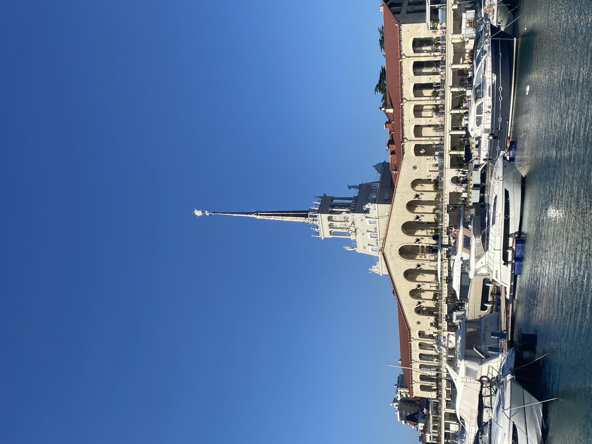The image captures a sunlit marina or port scene, likely the Sochi Marine Terminal, on a clear day. A grand, multi-story building with classical architecture, featuring numerous arched windows and balconies, stretches across the lower portion of the frame. The building has a light-colored facade and a reddish-brown roof. From its center, a tall, ornate spire, white with gold accents and topped by a star-like finial, rises dramatically towards the sky.
To the right of the building, several yachts and boats are docked in calm, blue-green water. One prominent dark-hulled yacht has the Cyrillic text "COKOЛ" (Sokol) and "VO" visible on its side. Further along the pier, a white yacht shows registration "AA 0897 RUS". A few small figures can be seen on the building's balconies and possibly on the boats, suggesting activity.
The time of day appears to be mid-morning or early afternoon, indicated by the bright, high sun and the distinct but not overly long shadows. The weather is perfectly clear with a vast, unbroken deep blue sky dominating the upper part of the image. Visible text on the building itself includes "ВОКЗАЛ" (VOKZAL, meaning "Station" or "Terminal") above an arch, and "COЧИ" (SOCHI) on an upper section, confirming the location as Sochi, Russia. The scene conveys a sense of serene maritime activity at a picturesque waterfront.
