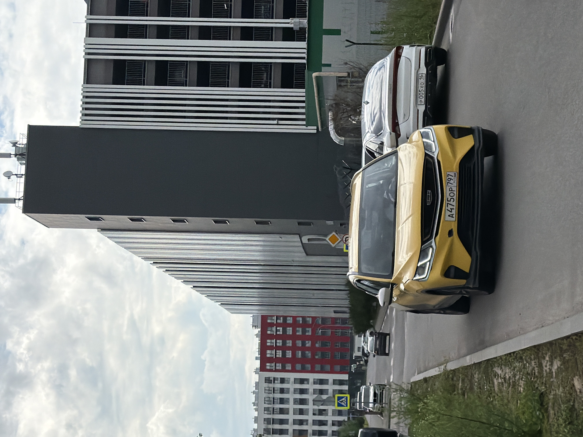 This media file depicts an outdoor urban scene with a focus on modern architecture and vehicles. In the foreground, a yellow car is parked on a paved surface, with its front end angled towards the viewer. Behind it, a white car is also parked. The cars are on a road or parking area.
The background is dominated by a large, multi-story building with a dark gray facade. Above this, a taller building with a white and black striped facade is visible, suggesting a contemporary architectural style. Further in the distance, other buildings, including one with a red facade and numerous windows, are present, indicating a densely built urban environment.
The sky is cloudy, suggesting overcast or partly cloudy weather. The time of day appears to be daytime. There are no people visible in the image.
Visible text includes license plates on the cars. The yellow car has a license plate that reads "A475OP 79Z". The white car has a license plate that reads "P 005XO 16". There are also road signs visible, one appearing to be a diamond-shaped warning sign and another a square blue sign with a white symbol.