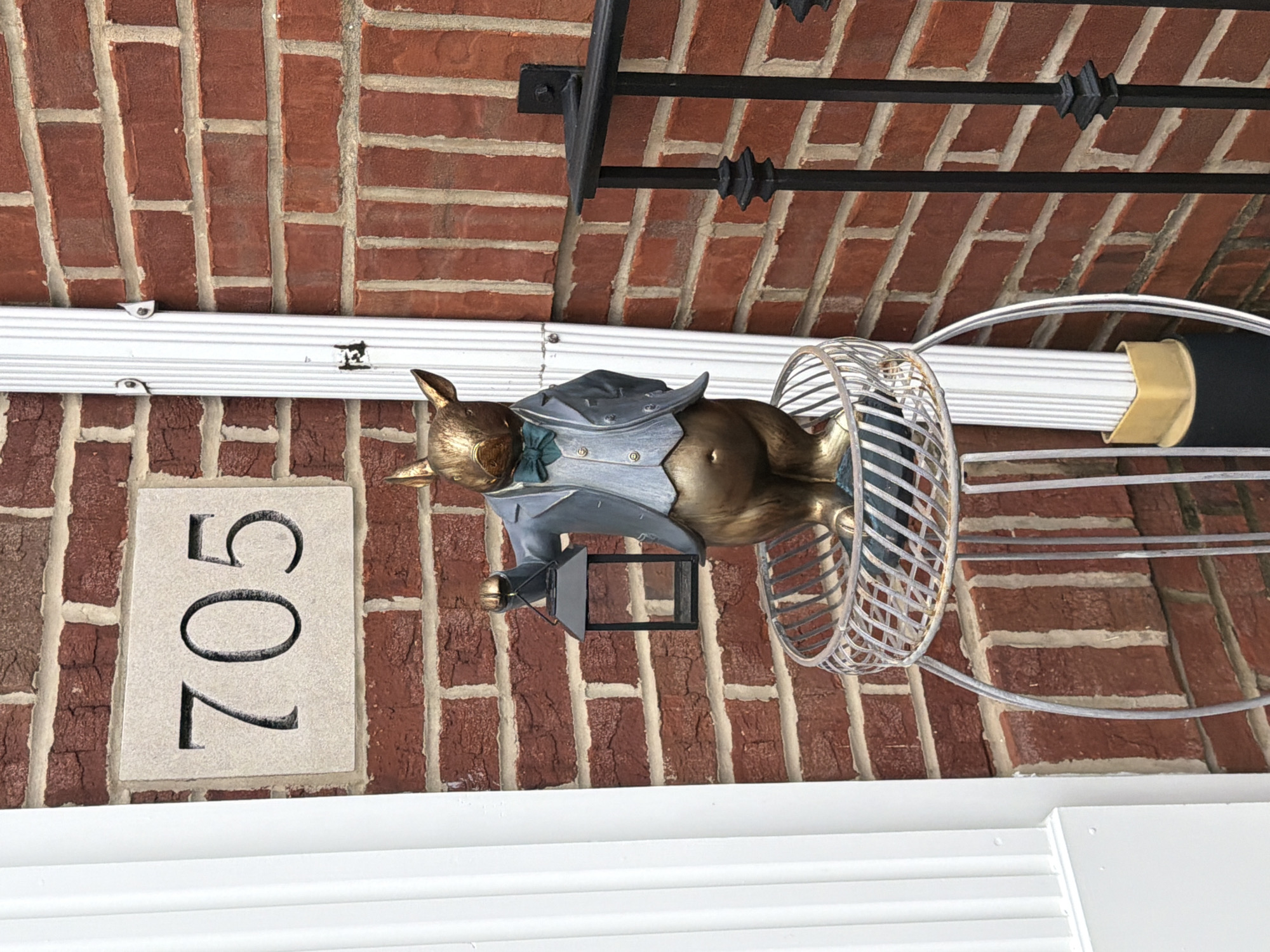 The image shows the exterior of a brick building with the house number "705" displayed on a stone plaque. A decorative statue of an animal, possibly a rabbit or a fox, dressed in a suit and holding a lantern, stands on a metal pedestal. A white downspout runs vertically along the brick wall next to the statue, and a black metal fence is visible on the right side of the image. The environment appears to be an outdoor residential setting.