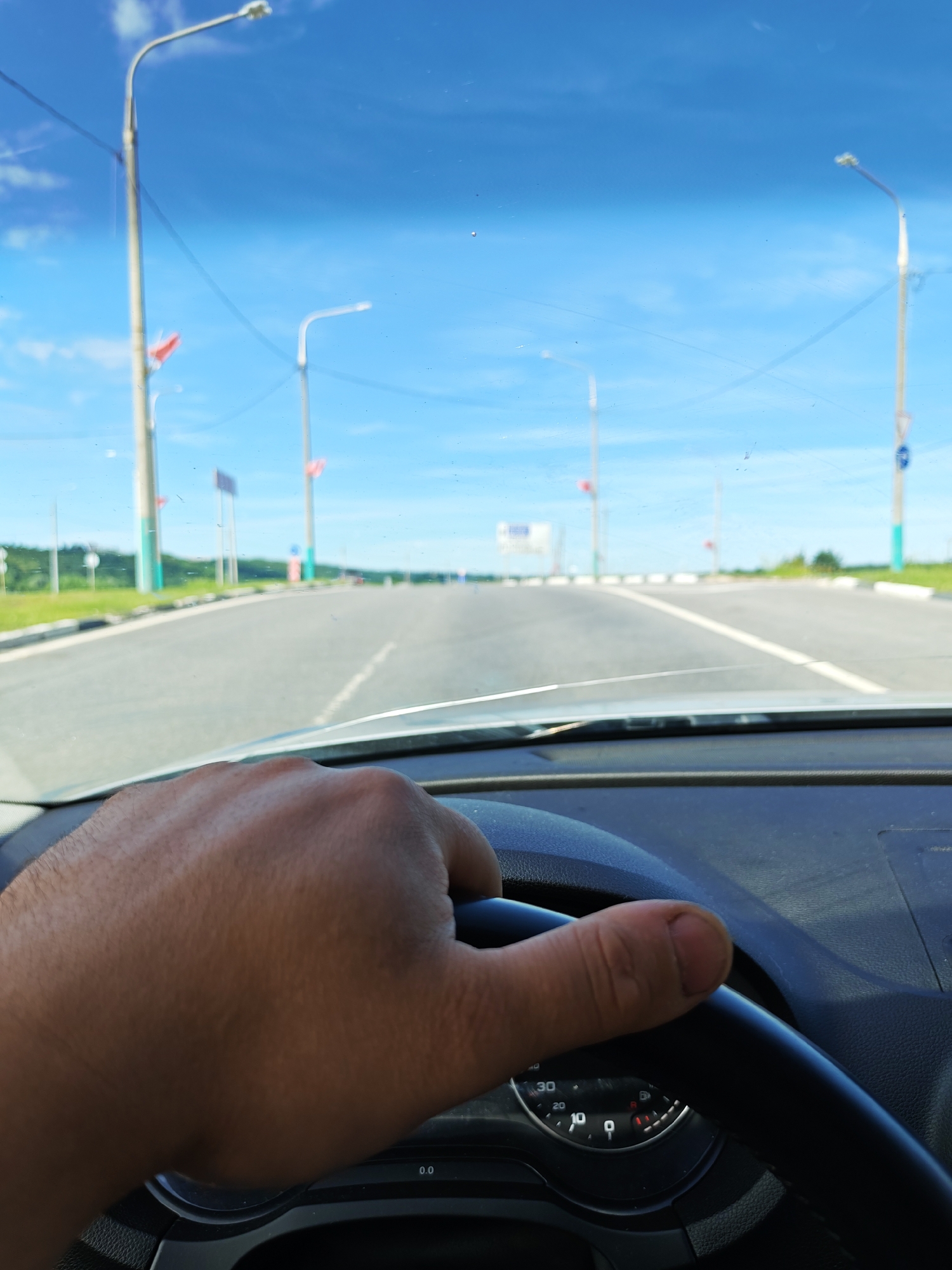 The media file depicts a first-person perspective from the driver's seat of a car, looking out through the windshield. The primary subject is the driver's hand, which is resting on the steering wheel, indicating the action of driving. The scene is an open road, possibly a highway or a wide, paved road with multiple lanes, stretching into the distance.
Several streetlights are visible along the sides of the road, suggesting an urban or suburban setting, or perhaps a well-maintained road in a rural area. Some of these streetlights have red flags attached to them, which could be decorative or part of a local event. In the distance, there are signs, indicating that the driver is on a route with traffic management. Greenery and trees can be seen bordering the road, interspersed with more poles and structures.
The sky is a clear, bright blue with a few scattered white clouds, suggesting it is daytime, likely midday or early afternoon given the brightness. The weather appears to be fair and sunny. There is no visible text in the image that can be clearly read, except for the "0.0" on a dial that is partially visible on the dashboard. The overall mood conveyed is one of travel and forward movement. The location context is identified as Kaluga, Russia, which aligns with the type of road infrastructure and the presence of flags that might be related to local traditions or events.