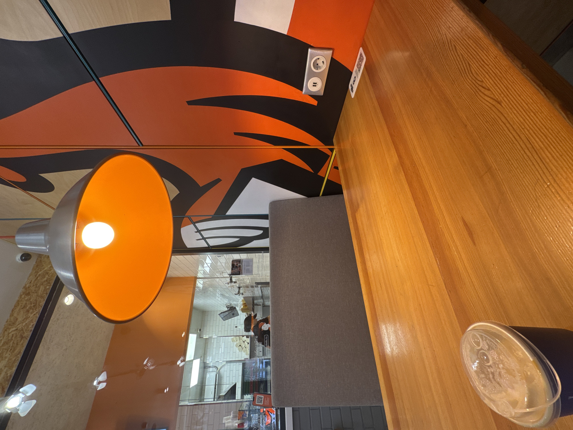 The image shows a modern interior, possibly a fast-food restaurant, in Rostov-on-Don, Russia. An orange pendant lamp is prominently featured in the foreground. Adjacent to the lamp is a wooden tabletop with a drinking cup on it. A grey cushioned bench is along the table. On the wall above the table, there's a decorative pattern in black, orange, and white, along with an electrical outlet and a QR code sign. Through a glass partition, the kitchen area is visible, with staff working and food preparation areas in the background.