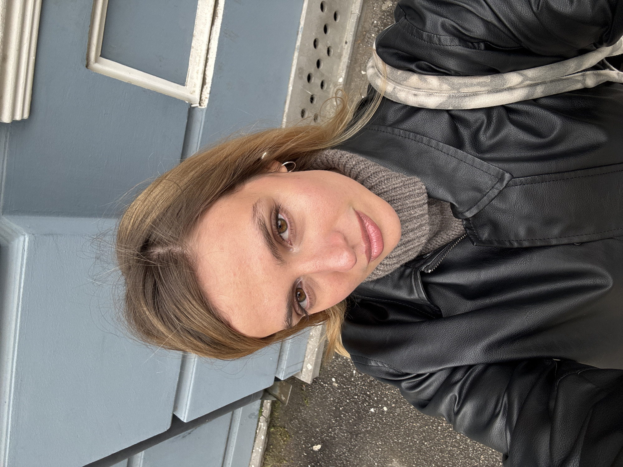 A medium close-up, high-angle portrait shows a young woman looking directly at the camera in an outdoor setting in Tbilisi, Georgia. She has light brown hair, styled with a side part, and warm brown eyes. Her fair complexion is accentuated by natural-looking makeup, including a pinkish lip color. She is dressed in a black faux leather jacket layered over a grey, ribbed turtleneck sweater, suggesting cool weather conditions. A small silver hoop earring is visible in her left ear. Over her right shoulder, a light-colored, possibly tie-dyed or patterned fabric strap, likely from a bag, is visible.
The background features a faded blue-grey building facade, appearing to be a door or wall, with white decorative trim and paneling that shows signs of wear. To the upper right, a grey metal grate or drain cover with circular perforations is partially visible, indicating urban infrastructure. The ground beneath is dark asphalt or concrete, appearing somewhat uneven and scattered with small debris, consistent with a sidewalk or outdoor pathway. The lighting is soft and diffused, suggesting an overcast day or indirect sunlight. The woman's expression is calm, with a slight, gentle smile. No visible text is present in the image.