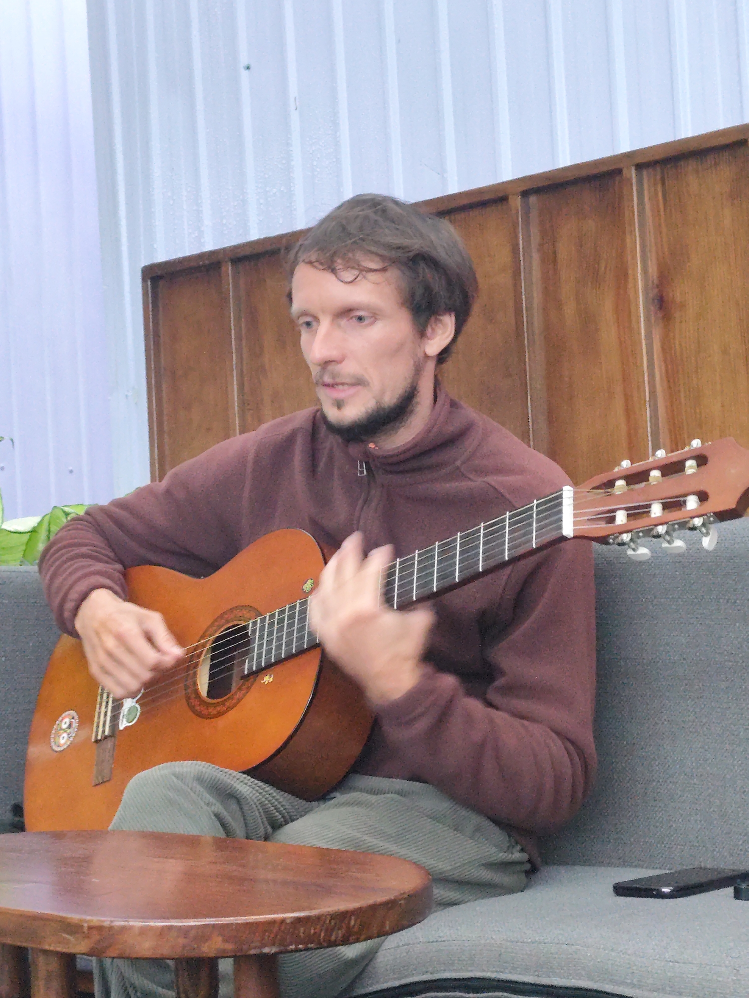 A man with light brown hair and a beard is sitting on a grey sofa and playing an acoustic guitar. He is wearing a maroon fleece jacket and light-colored corduroy pants. The guitar is brown with white tuning pegs. In front of him is a round, wooden table. Behind him, there is a wooden wall paneling and a white corrugated wall. A black smartphone is visible on the sofa to the right of the man. The lighting suggests an indoor setting, possibly a cafe or a residential space. This scene is not identifiable as being in Da Lat, Vietnam.