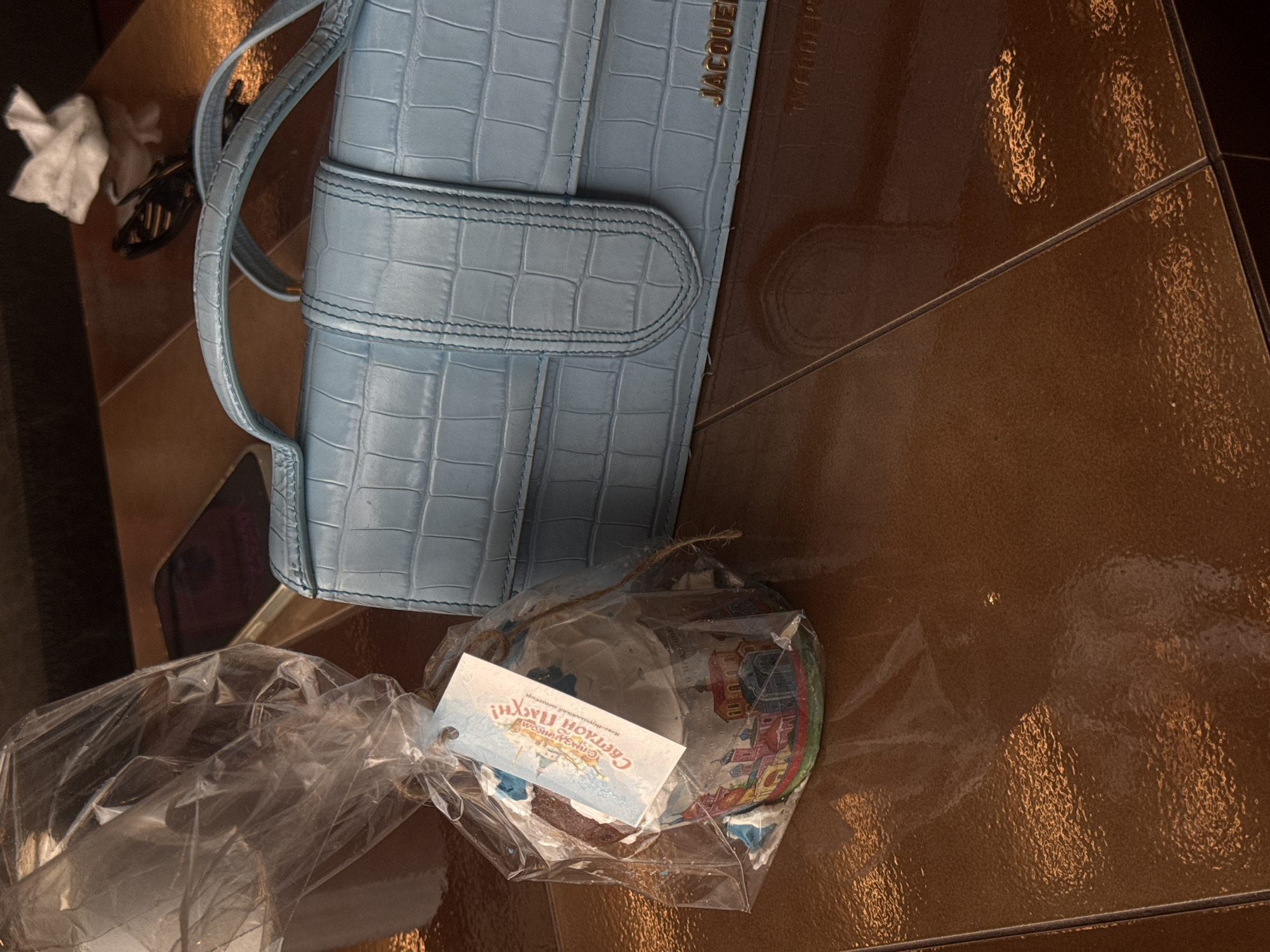 The image shows a light blue, crocodile-patterned handbag with the word "JACQUEMUS" visible on its side. Next to the handbag is a packaged item containing a decorated plate, tied with a string and a tag written in Russian. The tag mentions "СВЕТЛОЙ ПАХНИ!". The items rest on a brown, reflective surface, possibly a tiled floor or table. The location of Khimki, Russia is inferred from the Russian text on the tag. The scene suggests a recent purchase or gift related to a religious holiday.