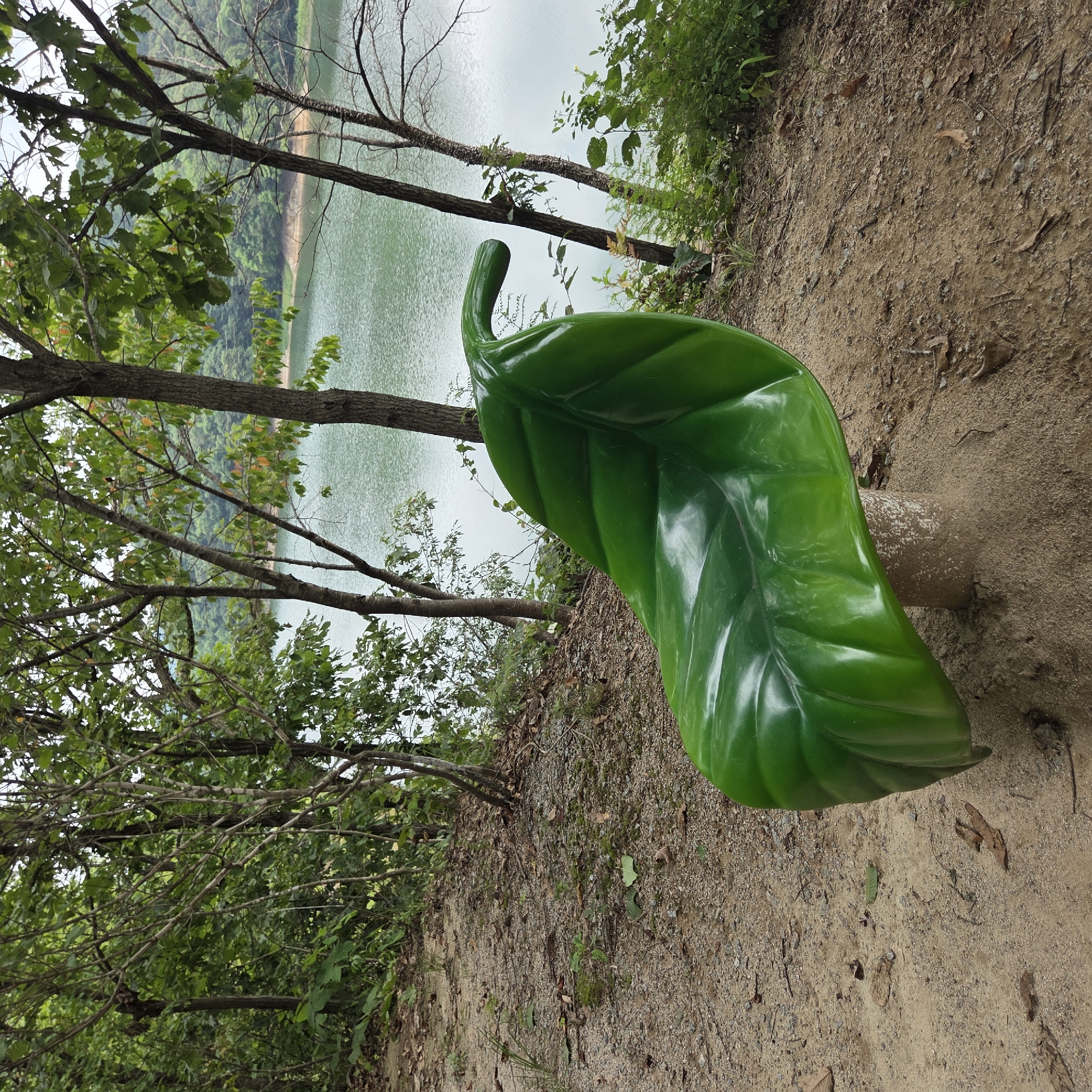 A close-up, eye-level shot captures a unique, bright green, leaf-shaped bench situated on a sandy, dirt path. The bench is prominently placed in the foreground, with its shiny, curved surface reflecting the ambient light. The seat of the bench is supported by a short, textured, cylindrical base.
Behind the bench, a tranquil lake with calm, greenish-blue water stretches into the distance. The lake is bordered by a densely wooded shoreline, with various trees and lush green foliage filling the background. Several tree trunks are visible, some slender and others thicker, interspersed with leafy branches that partially obscure the view of the water and the distant, hazy hills beyond. The ground leading up to the bench is a mix of sand, small pebbles, and fallen leaves, suggesting a natural, outdoor setting. The overall impression is one of a peaceful, natural park or recreational area, possibly in Gapcheon-myeon, South Korea, given the context. There are no people or animals visible in the frame.