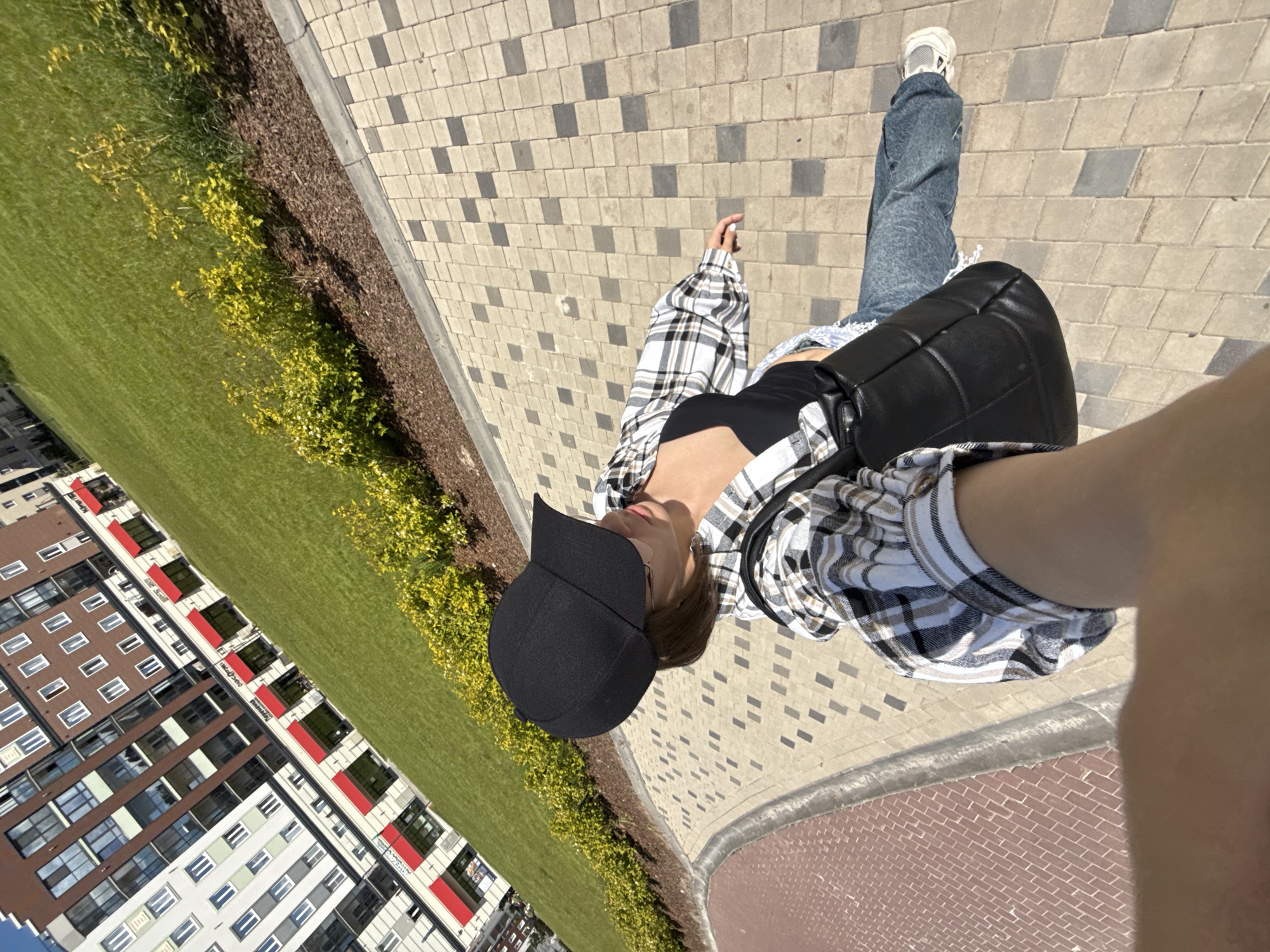 An overhead, high-angle outdoor shot captures a person and their surroundings in what appears to be a modern urban setting. The image is oriented with the ground extending diagonally from the bottom left to the top right.
The central subject is a person, seen from above and slightly to the front. They are wearing a black baseball cap, a black crop top, and a plaid button-up shirt in white, black, and beige tones, which is worn open. They have blue denim jeans and white sneakers. A black quilted shoulder bag is slung across their body. Their right arm, possibly holding the camera for a selfie, extends into the frame on the right.
The scene features a paved walkway made of light-colored interlocking pavers with a scattered pattern of darker grey squares. Adjacent to the walkway is a strip of dark brown mulch or soil, bordering a well-maintained lawn of vibrant green grass. Along the edge of the mulch and grass, there are several light green shrubs with yellowish leaves.
In the background, on the far side of the green space, stand several multi-story residential buildings with modern architecture. These buildings feature facades in various colors, including white, brown, and grey, with distinct red accents and numerous windows. Some red panels on the buildings contain small, unreadable white text, possibly indicating businesses or services like a "СПОРТИВНЫЙ КЛУБ" (Sports Club) or "ДЕТСКИЙ ЦЕНТР" (Children's Center) based on faint visual cues.
The weather appears sunny and clear, indicated by the bright lighting and distinct shadows. The overall impression is that of a pleasant daytime scene in a contemporary residential area, likely in Pervomaysky, Russia, as specified.
