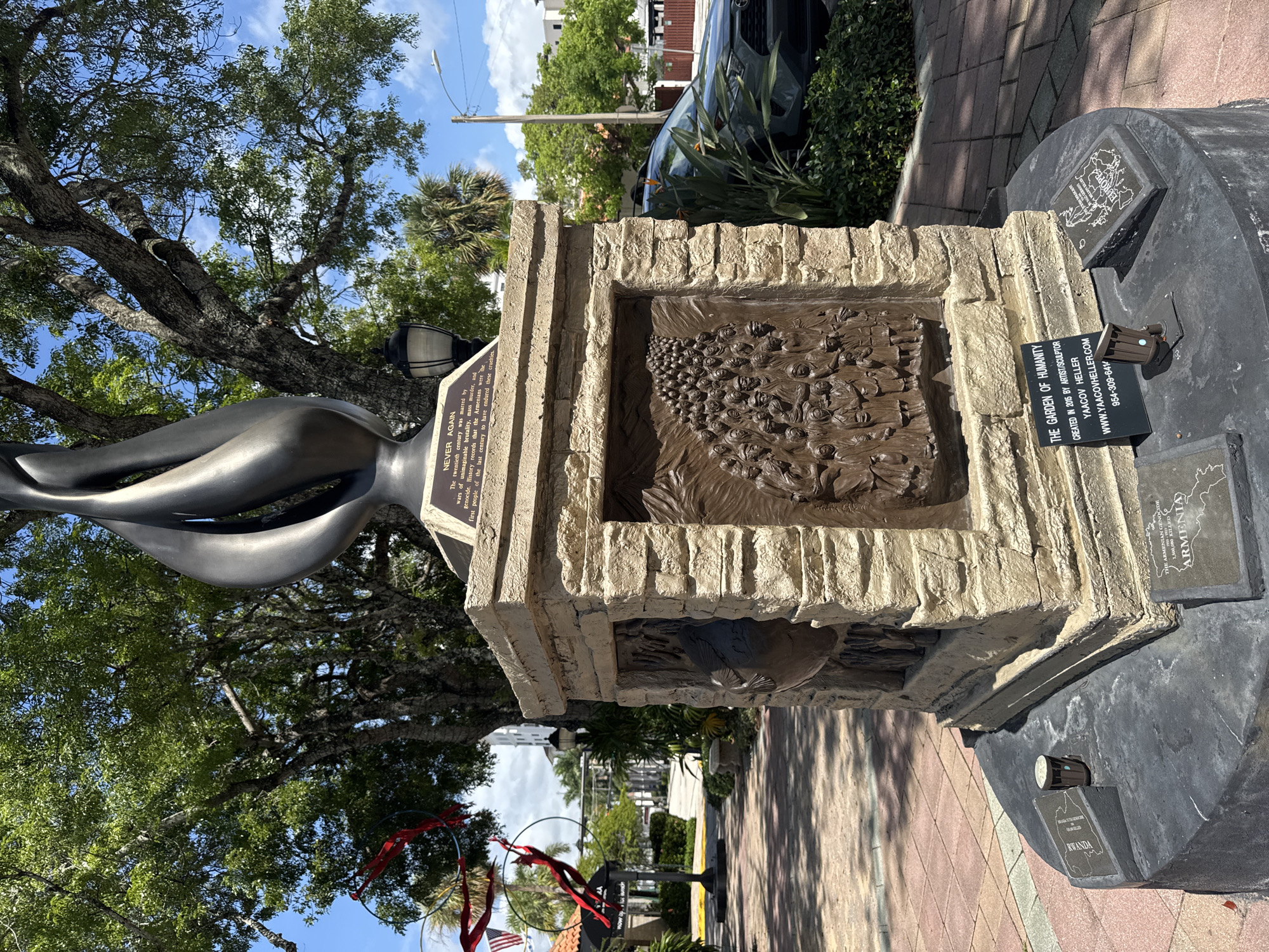 The image displays the "Garden of Humanity" memorial, located in Sanborn Square, Boca Raton, Florida, United States of America. The central feature is a monument consisting of a dark, abstract, flame-like sculpture surmounting a rectangular pedestal constructed from stacked, light brown, rough-hewn stone blocks.
On the front of the pedestal, a bronze relief depicts a dense crowd of figures, suggesting a multitude of people. To the left of this relief, another side of the pedestal features a different bronze relief, partially obscured, which appears to depict a dove or similar bird, possibly in flight or ascending.
Below the flame sculpture, a bronze plaque on the pedestal reads: "NEVER AGAIN. The twentieth century was marred by wars of unimaginable brutality, mass murders and genocide. History records that the Armenians were the first people of the last century to have endured these cruelties."
At the base of the monument, a circular, dark gray plinth supports several smaller, angled plaques with maps and text. Visible plaques include:
* One with the outline of Armenia and the text: "THE ARMENIAN GENOCIDE. 1,500,000 KILLED. ARMENIA."
* Another with the outline of Rwanda and text: "RWANDA. 1994. 800,000 KILLED."
* A third with the outline of Europe and the text: "THE HOLOCAUST. 6,000,000 KILLED. EUROPE."
* A vertical black plaque on the plinth identifies the memorial: "THE GARDEN OF HUMANITY. CREATED IN 2015 BY ARTIST/SCULPTOR YAACOV HELLER. WWW.YAACOVHELLER.COM 954-309-6419."
The monument is set on a patterned brick paver path. In the background, large, mature trees with dense green foliage dominate the upper portion of the frame under a bright blue sky with scattered white clouds. To the left, a contemporary art installation features several black rings from which red, flowing fabric streams downwards. Two small American flags are partially visible behind this installation. A dark-colored SUV is parked to the right of the monument, partially obscured by green shrubbery and a bird-of-paradise plant. A traditional black street lamp stands near the monument on the right. In the distance, several buildings and a utility pole are visible. The scene is brightly lit by natural sunlight, casting distinct shadows.