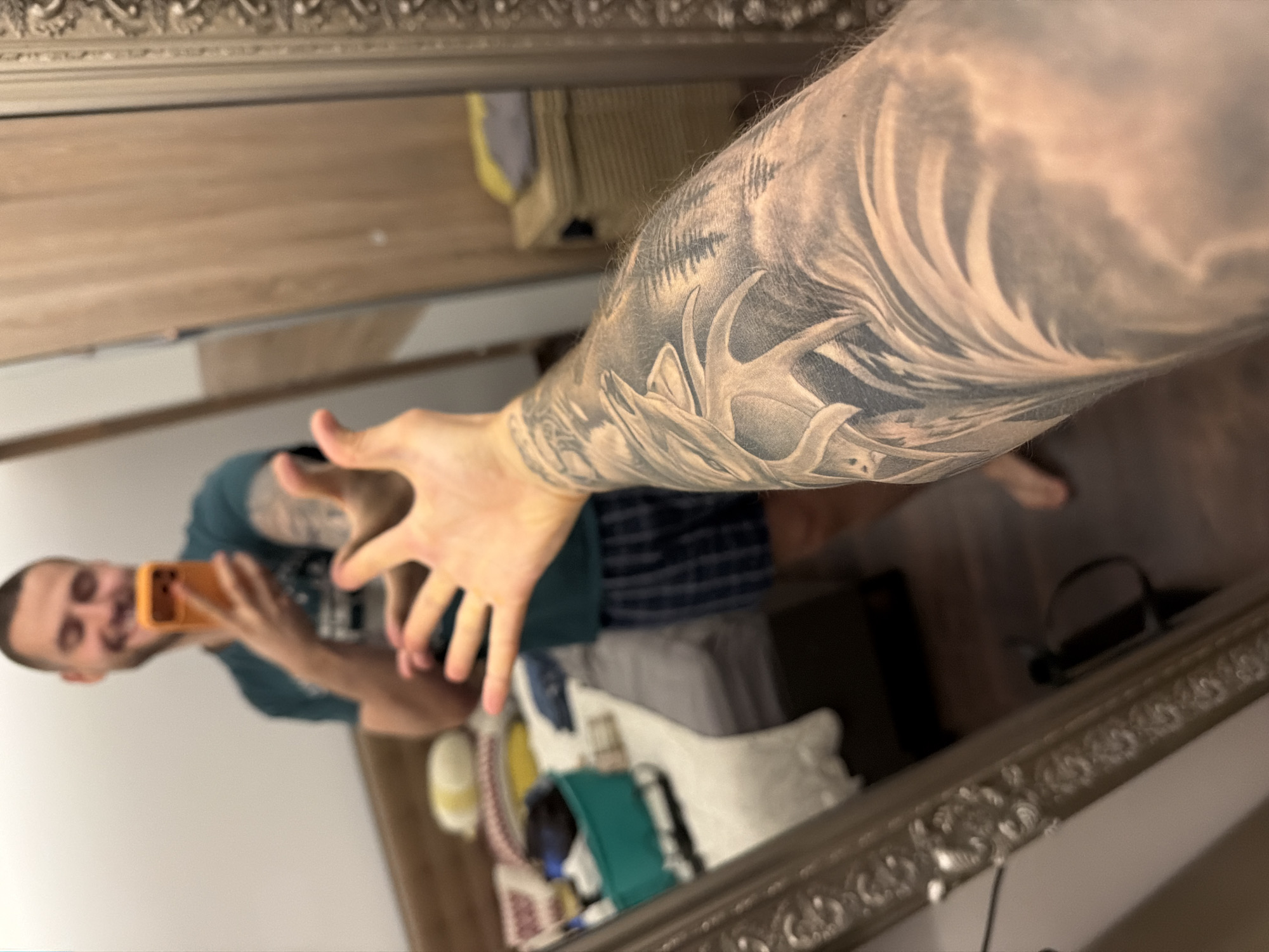The image is a first-person perspective shot of a man taking a selfie in a mirror. The man, who has short hair and a beard, is wearing a dark green t-shirt and plaid shorts. He is smiling and holding an orange smartphone to take the picture. His left arm is extended towards the camera, showing off a detailed tattoo of trees and possibly antlers. The background suggests a bedroom with a bed, a dresser, and a large, ornate mirror. The scene is indoors, likely in a residential setting. No specific location in Sofia, Bulgaria, can be determined from the visual information provided.