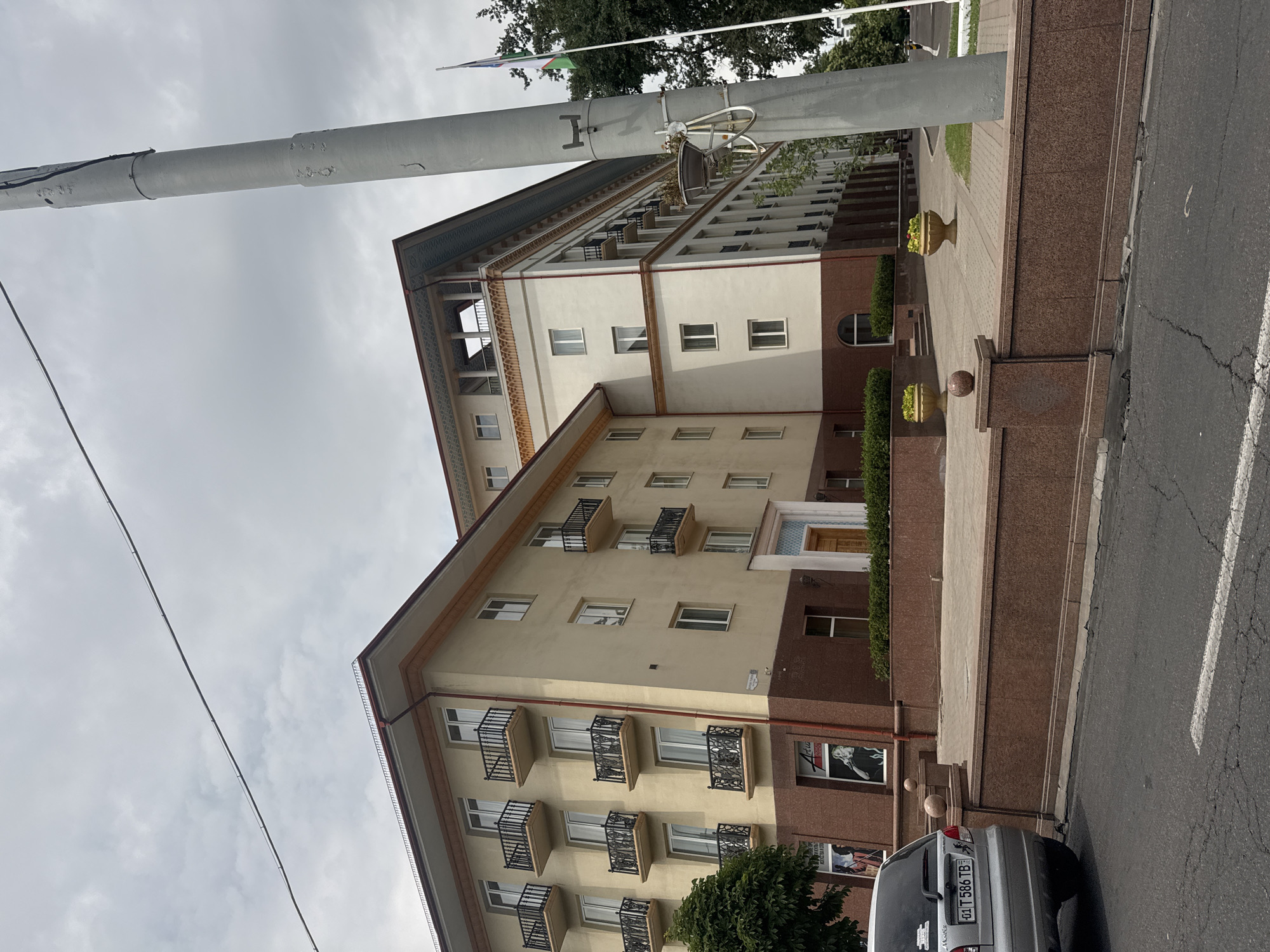 The image shows a wide, upward-angle shot of a building with a neoclassical design, likely in Tashkent, Uzbekistan. The building has a light-colored facade with a reddish-brown stone base. Multiple balconies with decorative black railings are visible along the facade. The sky is overcast, suggesting it's either early morning or late afternoon on a cloudy day. A large gray cylindrical pole, possibly a lamppost or utility pole, dominates the left side of the frame, obscuring part of the building and the sky. Trees can be seen at the top of the image. In the foreground, a paved walkway leads to the building, with manicured bushes and two decorative planters. A silver car with license plate "01 T 586 TB" is parked on the street to the right. Some posters or advertisements are visible on the lower part of the building's facade, one featuring a black and white image of a woman. The overall impression is an urban scene with a mix of architectural styles and everyday elements. There is no visible human activity in the frame.