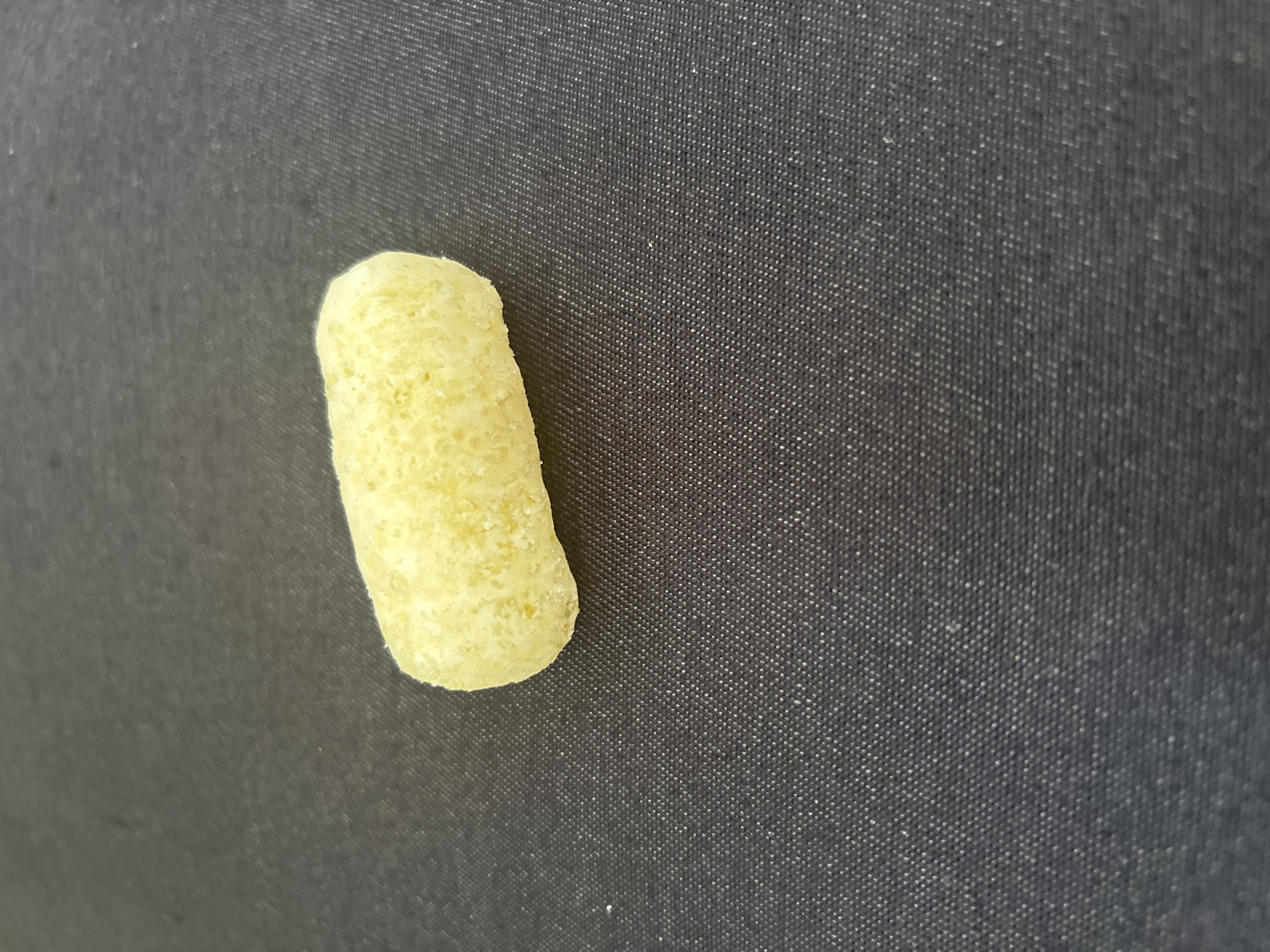 The image shows a single, pale yellow, oval-shaped puffed snack food item resting on a dark gray textured surface. The surface appears to be fabric with a visible weave pattern. The snack is centrally located in the upper half of the frame, with ample space around it. There are no people, actions, or interactions depicted. The lighting is even, with no harsh shadows. The background is out of focus, drawing attention to the snack.