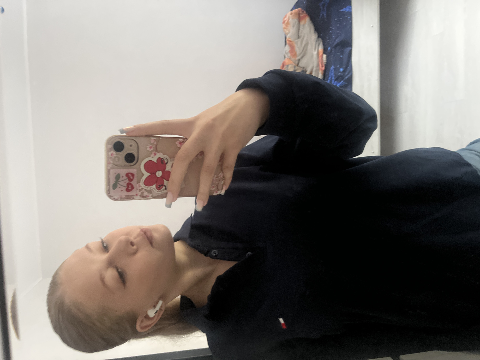 A young Caucasian female is taking a mirror selfie in a room. She is wearing a dark navy polo shirt with a small red, white, and blue logo on the left chest. She has white wireless earbuds in her ears and her hair is pulled back in a ponytail. She is holding a mobile phone in her right hand, which has a case decorated with cherries and a red flower. Her fingernails are painted a light grey color. The background shows a white wall and a glimpse of a bed with patterned bedding. The lighting is bright and appears to be from an overhead source. The scene is consistent with a bedroom or dressing area. There is no information in the image to suggest this is in the city of Perm, Russia.