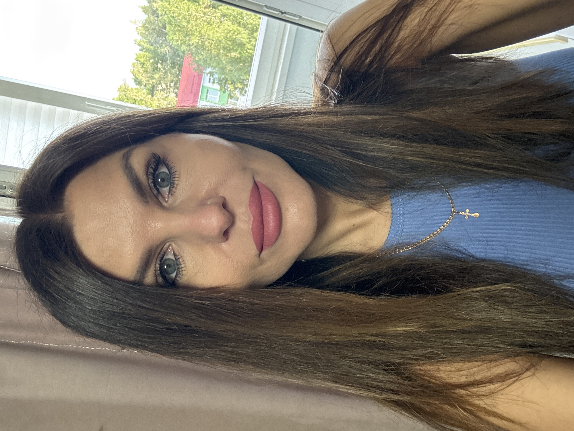 A medium shot selfie shows a woman from the chest up, looking directly at the camera. She has long, dark brown hair with lighter streaks, worn down. Her eyes are a light blue-green, and she is wearing subtle makeup, including pink lipstick. She is wearing a light blue, ribbed tank top with a delicate gold chain necklace featuring a small cross pendant. The background is split between a textured, light pink curtain on the left and a brightly lit window on the right, revealing trees and a building with a red roof outside. The scene appears to be indoors, possibly near a window overlooking a residential area in Chaikovsky, Russia.