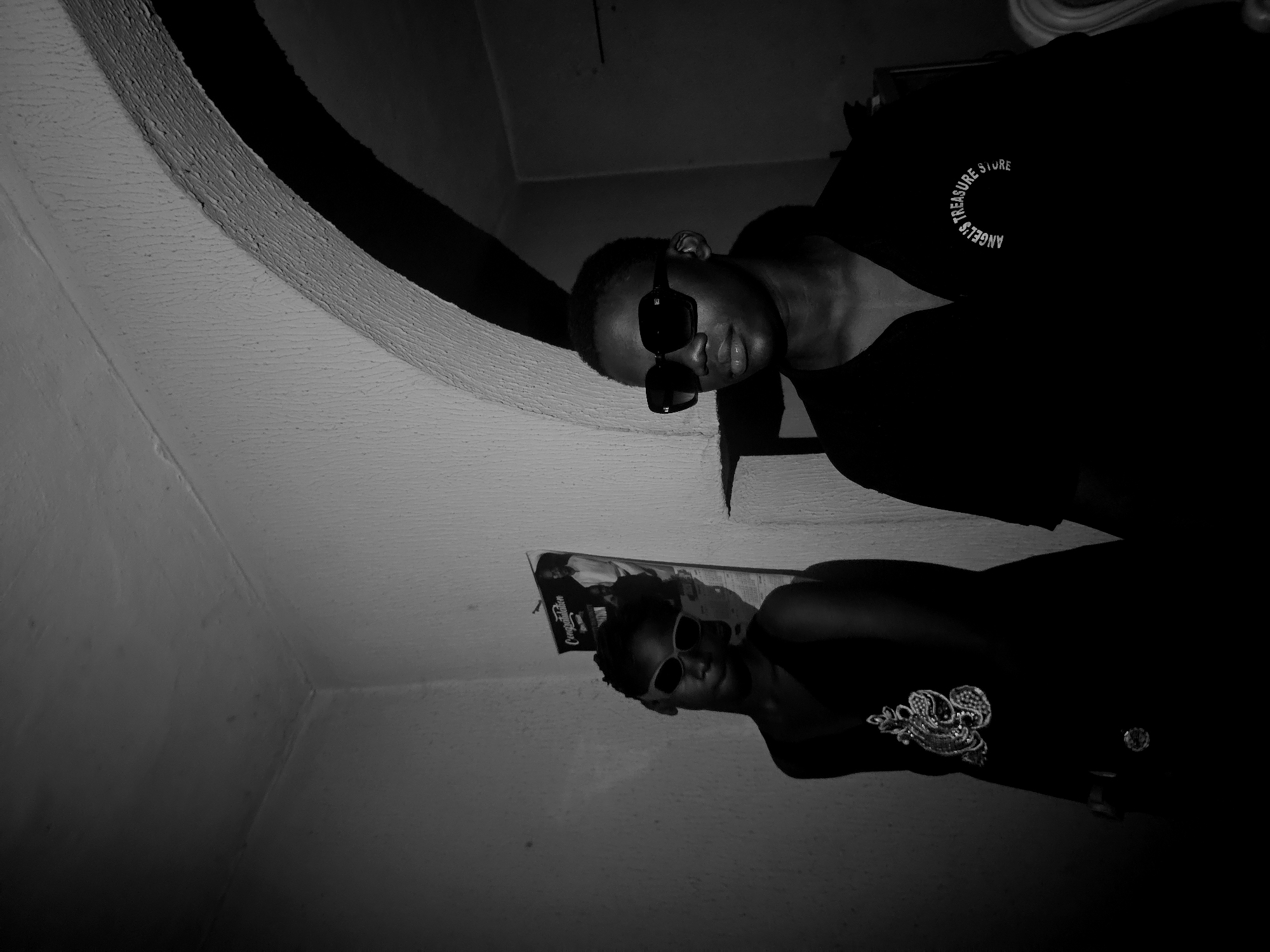 The black and white image presents an individual wearing sunglasses and a dark shirt with the inscription "ANGEL'S TREASURE STORE." A poster or magazine featuring a person is on the wall. The image is taken at an angle, with ceiling elements visible. The location is identified as Asaba, Nigeria.