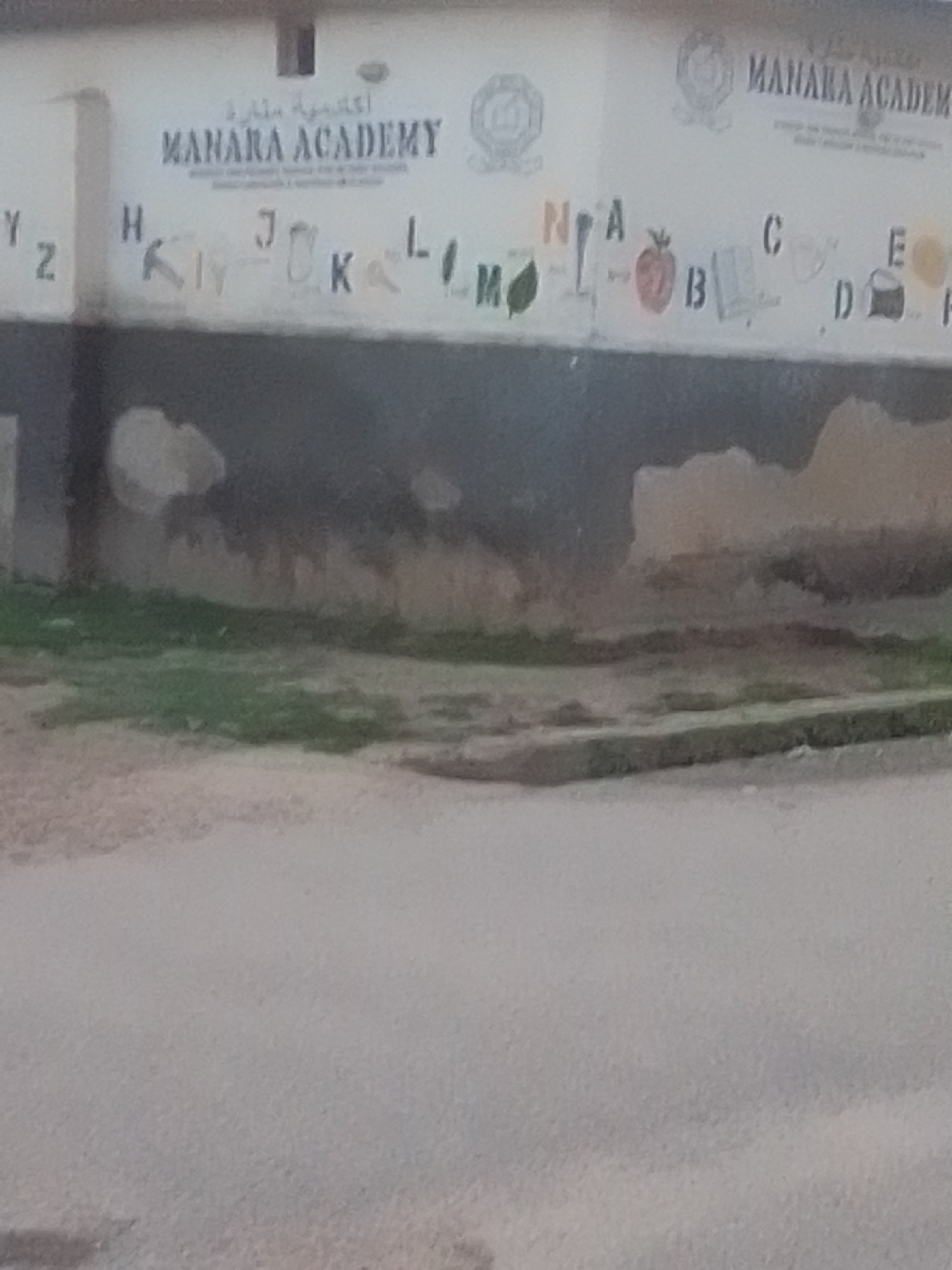 This is a medium shot of a mural on an exterior wall. The mural features the name "MANARA ACADEMY" prominently, with Arabic script above it. Below this, various letters of the alphabet are displayed, some with accompanying illustrations. For example, "J" is next to an illustration of a key, "K" appears to be next to some handwritten markings, "L" is followed by a leaf, and "M" is also next to a leaf. Further along, "N" is next to what looks like a spoon, "A" is next to a tomato, "B" is next to an open book, "C" is next to a circle, "D" is next to a drawing of a coconut, and "E" is next to a drawing of a small object. To the far left, the letters "Y" and "Z" are visible. The wall itself is painted a light color and appears to be weathered, with some patches of peeling paint and darker marks. The lower portion of the image shows a grassy area in front of the wall, and beyond that, a paved surface. The location is stated to be Liji Hill, Nigeria.