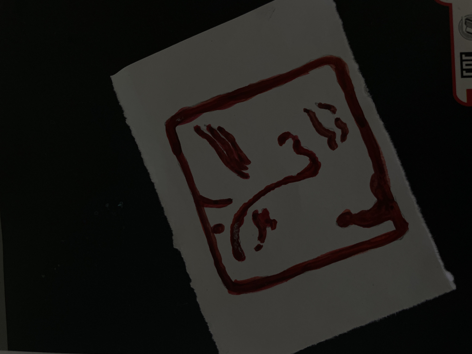 A close-up, low-angle shot shows a torn piece of white paper on a dark, out-of-focus background. A red ink drawing of a face is centered on the paper, enclosed within a square outline. The face is stylized, with prominent eyes and lips. A small, silhouetted figure appears to be kneeling or bowing at the bottom left of the face. The lighting is dim, with a spotlight effect illuminating the drawing. There is no discernible context or specific location identifiable within the image.