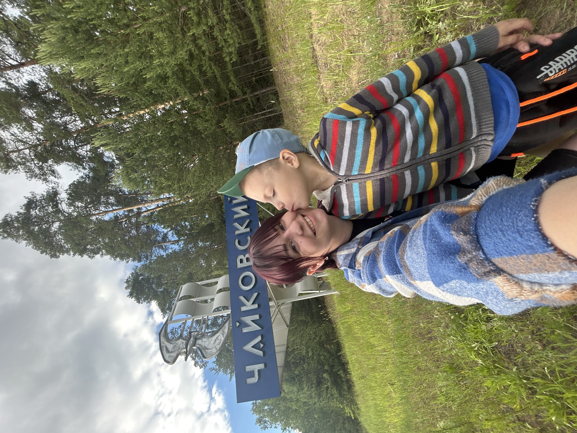 In the city of Chaikovsky, Russia, a young boy wearing a blue cap and a striped jacket kisses a woman on the cheek. They are positioned in front of a blue sign that reads "ЧАЙКОВСКИЙ" in white Cyrillic letters, with an abstract sculpture above it. The scene is set in a grassy area with tall pine trees in the background, under a partly cloudy sky.