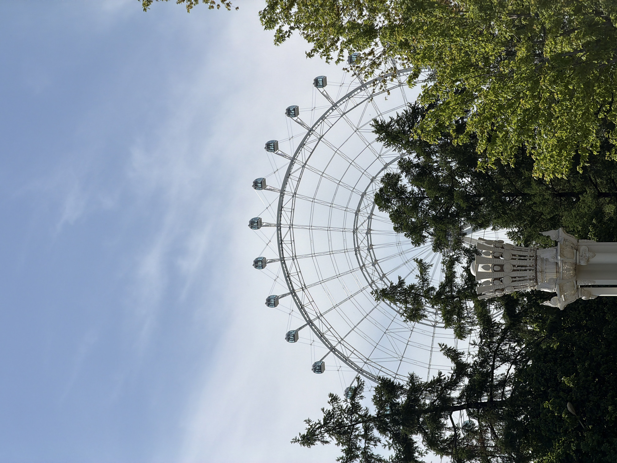 The image shows a Ferris wheel against a blue sky. The wheel is predominantly white with enclosed passenger cars. Green trees frame the right side and top of the wheel. The base of the wheel appears ornate and white. The location is Moscow, Russia.