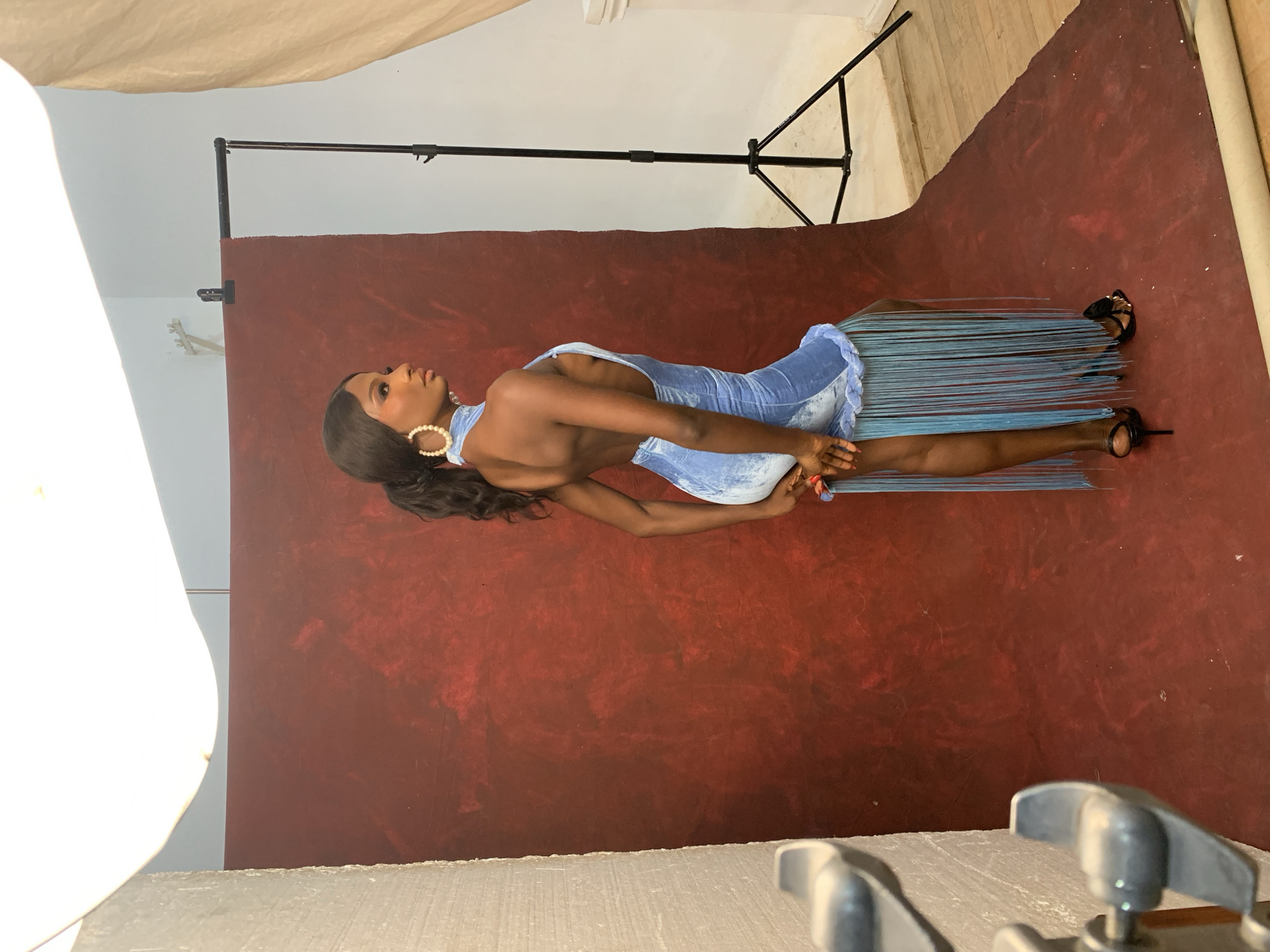 A young Black woman with dark skin and long, dark hair in a ponytail stands in a studio setting. She is wearing a light blue, backless, fringed dress and black high-heeled sandals. The woman is looking to her right, with her left arm bent and resting on her hip, and her right hand touching her left leg. She has large hoop earrings with pearl-like beads.
The background consists of a mottled dark red backdrop, with a white wall and some studio lighting equipment visible on the left. A black tripod with a light stand is positioned behind the woman to the right, and a beige drape hangs on the far right. The floor appears to be made of light-colored wood. The scene is indoors. The question about the specific location in Jos, Nigeria, cannot be answered from the provided image.