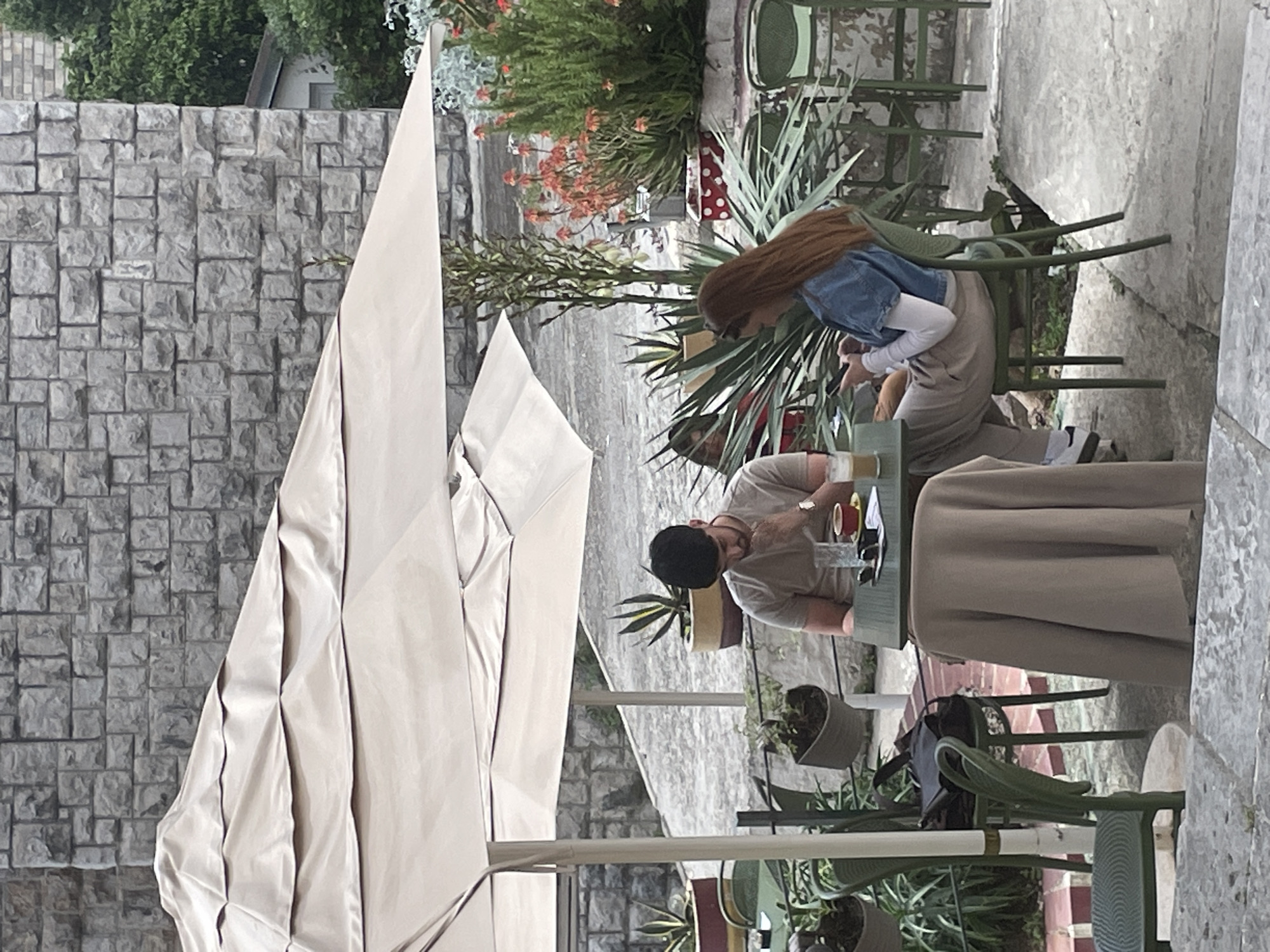 This image captures an outdoor cafe scene in what appears to be Herceg Novi, Montenegro, depicting two individuals seated at a table under a large, light-colored umbrella.
On the right side of the frame, a person with long, reddish-brown hair is seated, wearing a denim jacket over a white long-sleeved top and light-colored trousers, with what seem to be white sandals. They are leaning forward, looking down, possibly at a phone or something on the table. Opposite them, a person with dark hair wearing a cap and a light-colored t-shirt is also seated, engaged in a similar activity, looking down at the table. On their table, there are drinks, including an iced beverage in a tall glass, and what might be small food items. A beige tablecloth covers part of the table.
The setting is characterized by a grey stone wall on the left side, composed of rectangular blocks. Behind the seated individuals, lush green foliage and flowering plants, including spiky agave-like plants and a potted plant with red and white polka dots, create a vibrant backdrop. Green metal chairs and tables are scattered around, with a black bag resting on one of the chairs. The ground appears to be a light-colored, possibly concrete or stone, surface.
The activity suggests a casual meal or coffee break at an outdoor establishment. The lighting indicates daytime, but the presence of the large umbrella suggests a need for shade, implying a mild to warm day, not necessarily direct harsh sunlight. There is no visible text in the image. The overall mood is relaxed and peaceful.