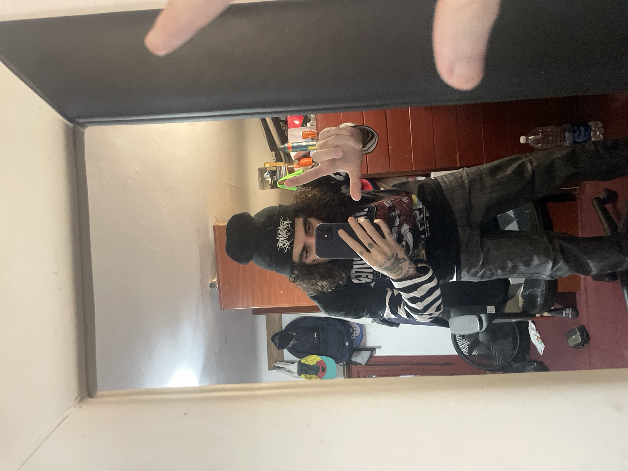 A person with long, dark, curly hair and wearing a black beanie, a striped long-sleeved shirt, a black graphic t-shirt, and dark pants is taking a selfie in a mirror. The person has tattoos on their arms and is making a hand gesture. The background includes a dark brown cabinet, a water bottle, a fan, and various items on a shelf. A backpack is visible hanging on a wall. The location appears to be a room within a residence. There is no information to definitively determine that the image was taken in Buenos Aires, Argentina.