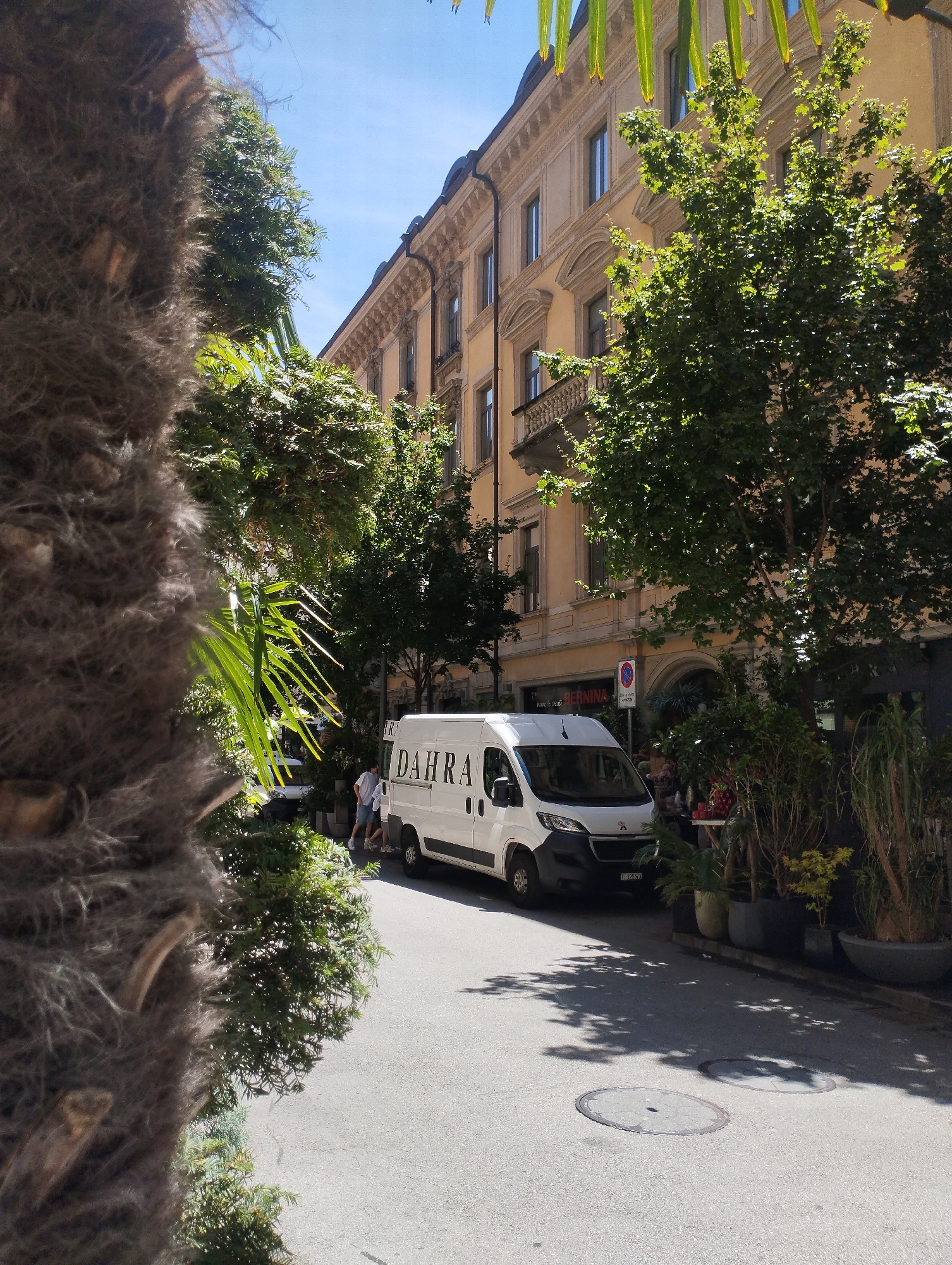 A white delivery van with the logo "DAHRA" is parked on the street in front of a building. The van is facing to the right and has its rear doors slightly ajar. Two people, a man and a woman, are standing on the sidewalk next to the van, appearing to be involved in unloading or loading the vehicle. They are casually dressed. The street is paved with asphalt and has manhole covers. Trees and potted plants line the sidewalk, providing shade. The building behind them is multi-story with a light yellow facade and ornate architectural details. The sky above is blue with some scattered sunlight filtering through the leaves of the trees. The overall impression is of a typical urban street scene on a sunny day in Lugano, Switzerland, with the presence of businesses and residential buildings.