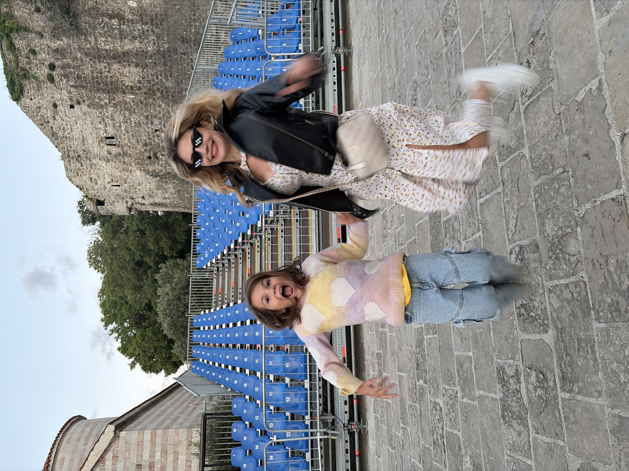Two females are captured in a dynamic pose, seemingly mid-jump, against the backdrop of an outdoor seating arrangement, a stone pathway, and architectural structures. The setting is suggestive of Budva, Montenegro, given the distinct stone structures.
The older female, dressed in a light-colored floral dress and a black leather jacket, accessorized with sunglasses and a crossbody bag, is positioned above, exhibiting a sense of motion. The younger female, clad in a patterned sweater and jeans, is below, mouth open in a possible expression of excitement or surprise.
Behind them, rows of blue chairs constitute an outdoor seating area, with metallic framework visible. Adjacent to the seating, a stone pathway extends, adding texture to the foreground. In the background, a stone wall and a building structure indicate local architectural features, typical of old Budva, Montenegro. The sky is overcast, suggesting diffused lighting in the scene.