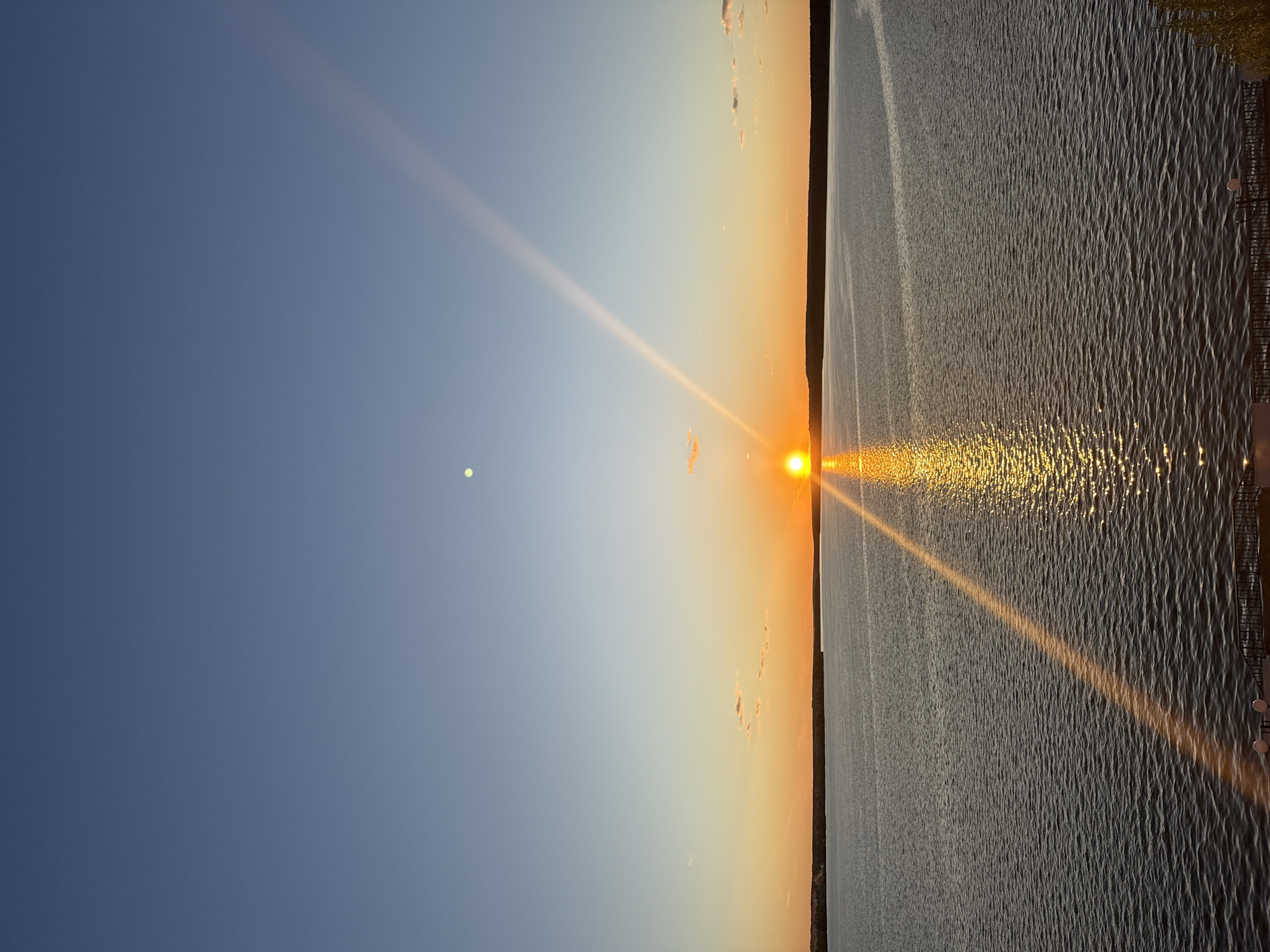 The image depicts a sunset over water, likely a body of water near Votkinsk, Russia, given the request for location specificity. The sun is positioned near the horizon, creating a bright reflection on the water's surface. A linear sunbeam is visible across the water and into the sky. The sky transitions from a deep blue to a warmer orange and yellow near the horizon. There is a small, round object in the sky, possibly the moon. The water has a textured appearance, indicating ripples or small waves. Along the horizon line, there is a dark strip of land or vegetation. A portion of a fence is visible on the right side of the image.