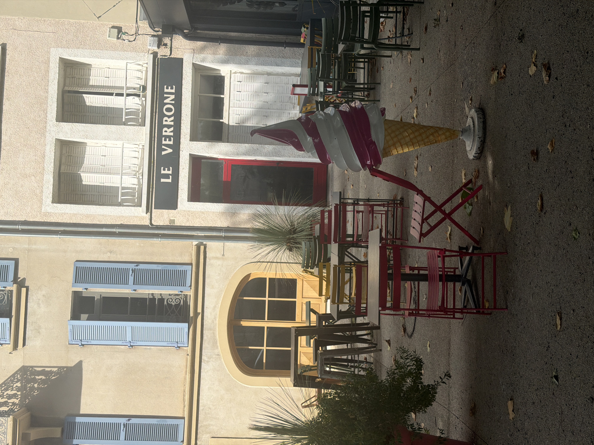 The image depicts an outdoor seating area in front of buildings. A prominent feature is a large, decorative ice cream cone statue with pink and white swirls, placed on a circular base. In the foreground, several red metal chairs and tables are arranged. Further back, more tables and chairs, some of them green, are visible. The ground is paved with asphalt and scattered with fallen autumn leaves.

The buildings in the background have a light-colored facade. The building on the left has large arched windows with wooden frames and light blue shutters. The building on the right features two upper windows with white shutters and balconies, and a dark sign above a red door that reads "LE VERRONE". This sign suggests the establishment might be a restaurant or cafe. The presence of outdoor seating and the name of the establishment imply this is likely a commercial space within a town or city.

While the exact location within Mercurol-Veaunes is not specified by any visible landmarks, the architectural style and the overall ambiance are consistent with a European town setting. The scene is tranquil, with no people present, and bathed in sunlight, casting shadows from the buildings and furniture. The lighting indicates it is daytime.