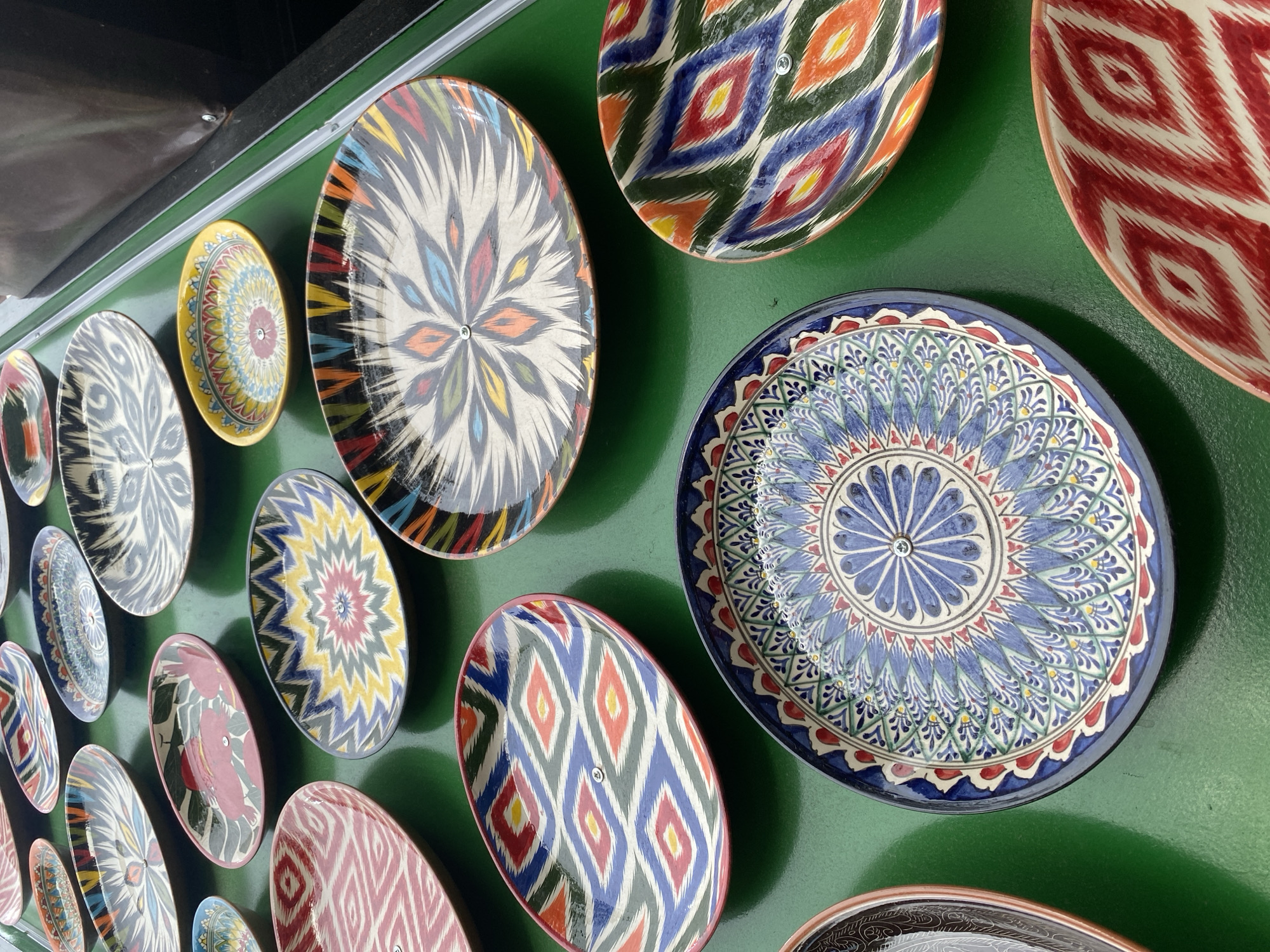 An overhead, medium-shot captures a collection of vibrantly patterned, oval-shaped plates arranged on a dark green, reflective surface. The plates, likely ceramic, display intricate geometric and floral designs in a variety of color combinations, including red, white, blue, yellow, green, and black. Some plates feature a central rivet, suggesting they were once part of a larger display or structure. The lighting is bright and even, casting soft shadows and highlighting the glossy finish of the plates. The arrangement of the plates suggests a market or artisan display, possibly in Almaty, Kazakhstan, given the style of the patterns which are reminiscent of traditional Central Asian textiles. The background is blurred, with a dark, out-of-focus area suggesting a stall or tent, and a sliver of a light-colored wall or ceiling. There are no people or animals visible in the frame.