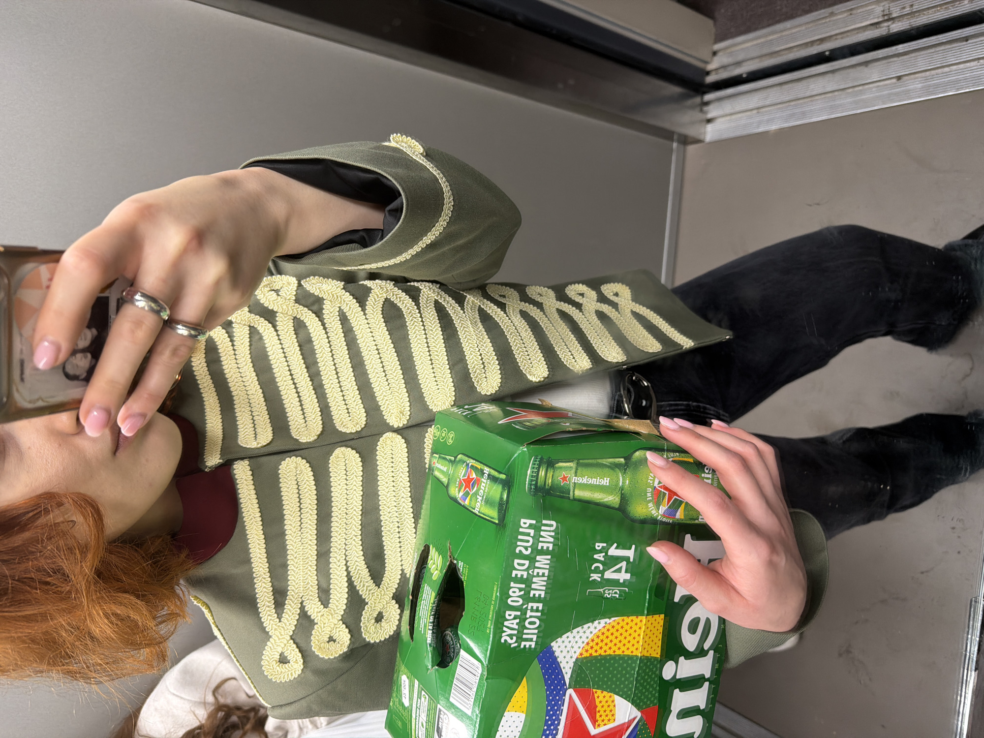 A person is holding a 14-pack of Heineken beer and a smartphone. The person is wearing a military-style jacket with gold trim and black jeans. The scene appears to be an elevator. No specific location in Strasbourg, France, is visible.
