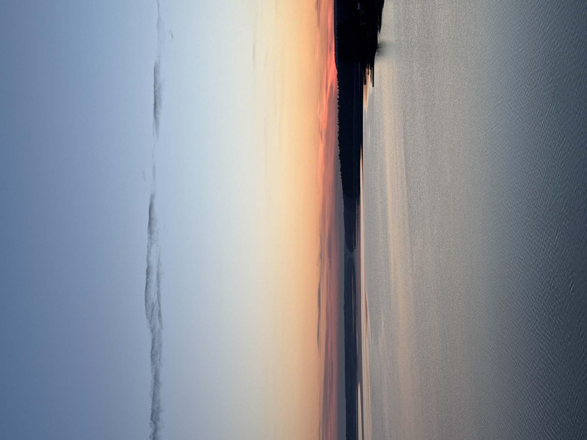 The image displays a serene twilight scene over a large body of water, likely the Votkinsk Reservoir in Russia. The sky, occupying the upper half of the frame, transitions from a clear, pale blue on the left to warmer shades of soft orange, yellow, and subtle pink closer to the horizon on the right. A long, thin, wispy cloud stretches horizontally across the cooler blue portion of the sky.
Along the horizon, a dark, undulating silhouette of a distant landmass, covered in dense trees, separates the sky from the water. A few faint, indistinct dark shapes, possibly distant structures or navigation markers, are barely discernible on the far right of this treeline.
The lower half of the image is dominated by the calm surface of the water, which reflects the soft, diffused light of the sky. The water exhibits a textured appearance with subtle ripples, appearing in muted tones of blue, grey, and gentle hints of the sky's warmer colors. The soft lighting suggests either a sunrise or sunset, creating a peaceful and contemplative atmosphere. No specific activity or visible text is present.