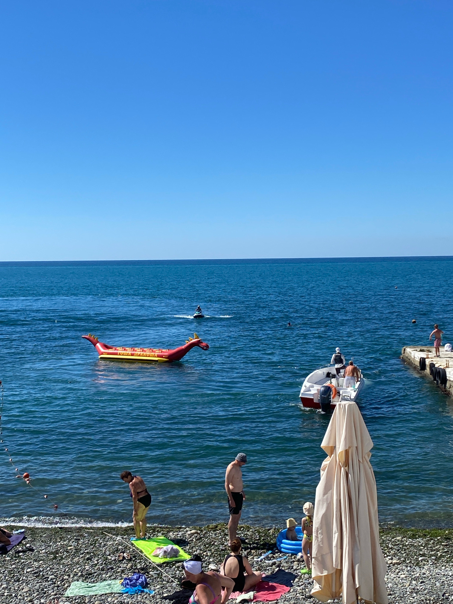 A bright, sunny day scene unfolds at a pebbled beach in Sochi, Russia, with the deep blue Black Sea as the backdrop. The water is calm, dotted with various vessels and people enjoying aquatic activities. In the mid-ground, a red inflatable "dragon" watercraft, bearing Russian text, floats on the water. To its right, a white motorboat with several people aboard is heading towards a concrete pier. A person on a jet ski is further out in the water. Closer to the shore, several individuals are visible on the beach. A woman in a black bikini stands on a lime green towel near the water's edge, looking down. A man in black shorts stands nearby. In the foreground, a large, cream-colored beach umbrella is folded and stands prominently on the pebbles. Other beachgoers are seated or lying on towels, and a small inflatable pool with two children is also present on the beach. The clear blue sky indicates fair weather.