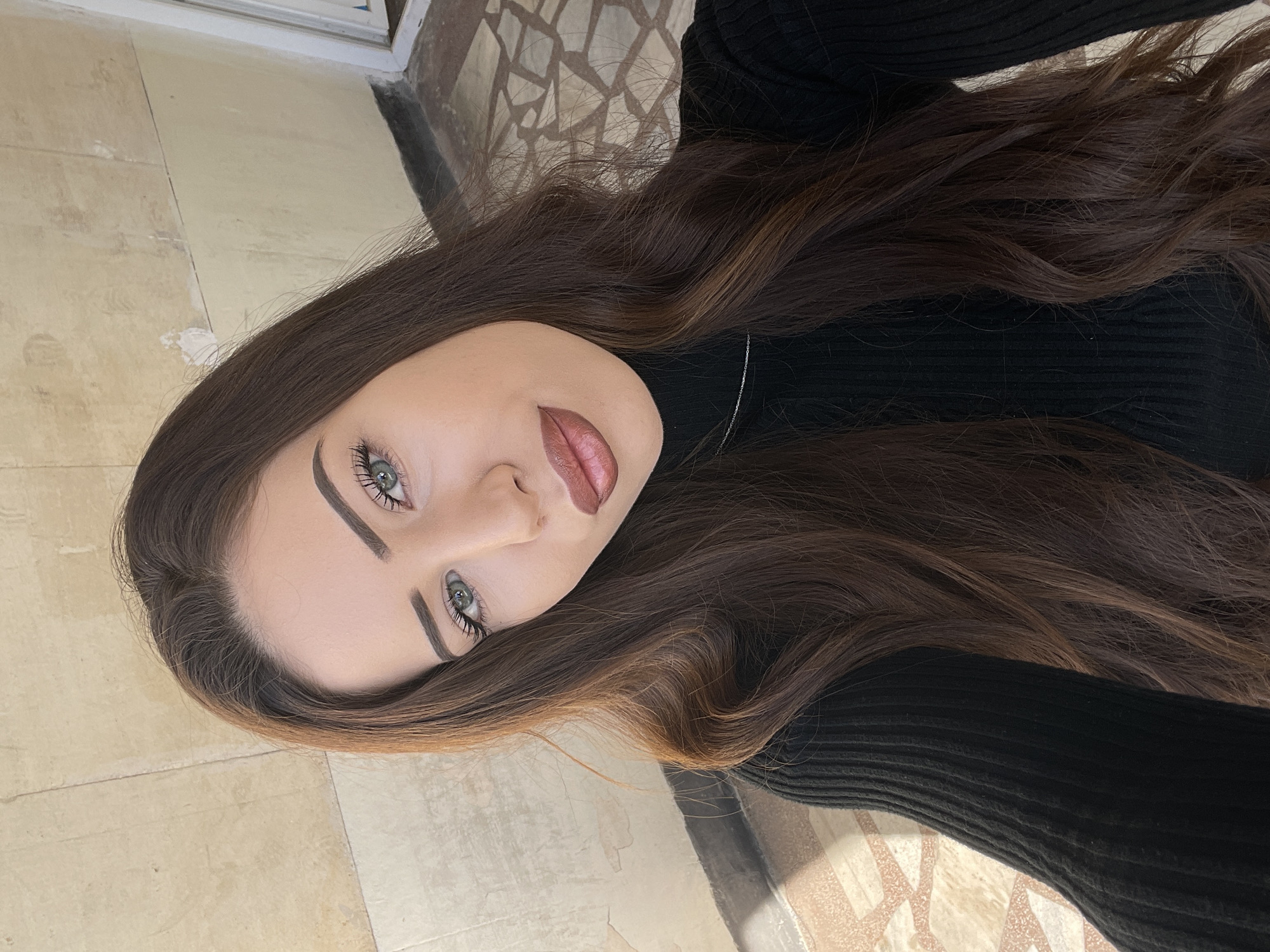 A close-up, rotated perspective shot of a young woman with long, brown wavy hair and fair skin. She has light-colored eyes, defined eyebrows, and is wearing lip makeup. The woman is wearing a black turtleneck sweater and a thin silver necklace is visible. The location is suggested to be in the city of Veliko Tarnovo, Bulgaria, based on the presence of tiled flooring. The floor is beige and light brown near the subject but turns to complex geometrical shapes made of marble in the upper right part of the image. It's impossible to confirm the location based only on the image.