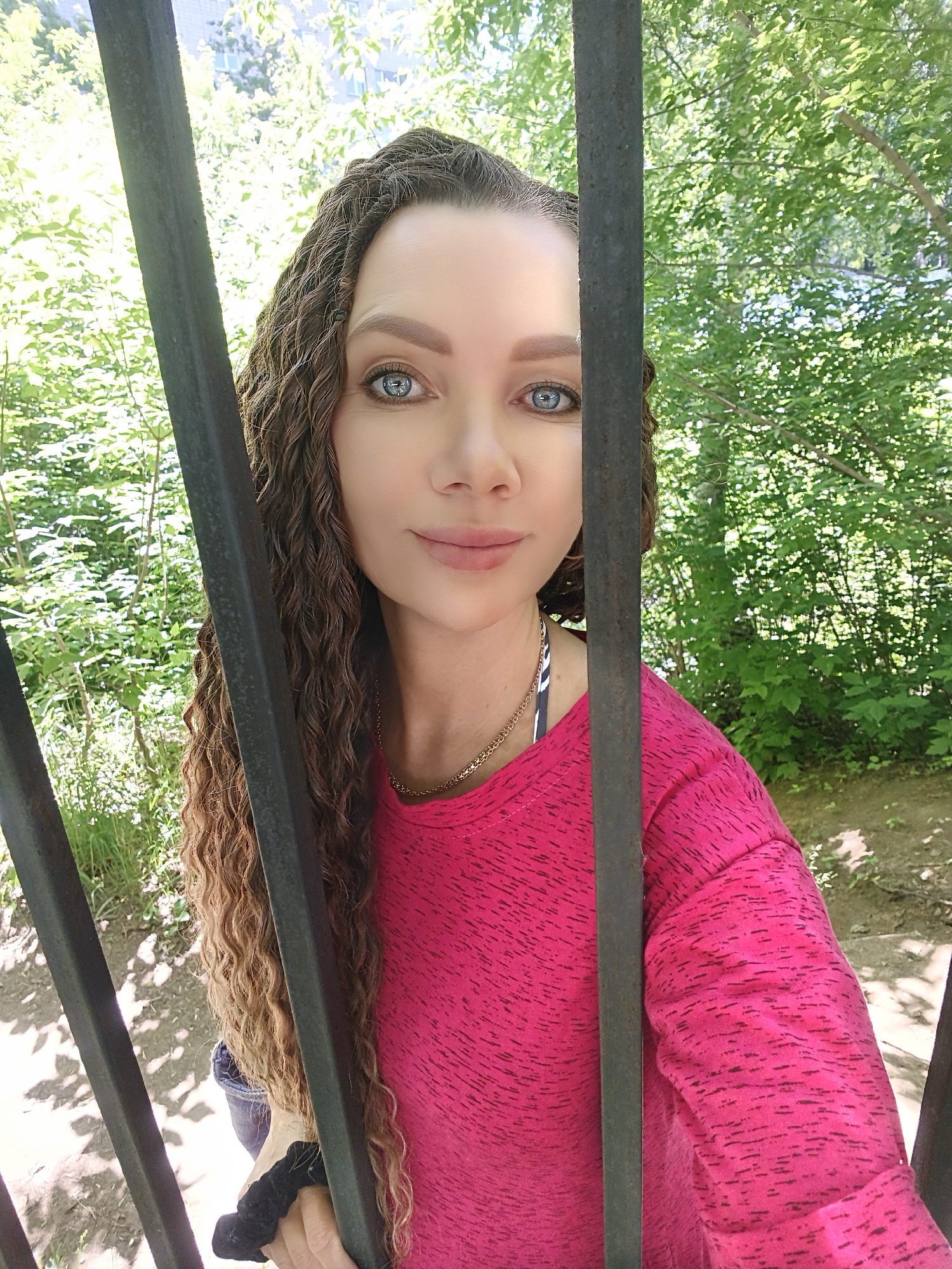 A woman with long, curly, blonde hair and blue eyes is standing behind a black metal fence in an outdoor setting. She is wearing a pink long-sleeved shirt and a gold chain necklace. The background consists of lush green trees and foliage. The lighting suggests it is daytime.