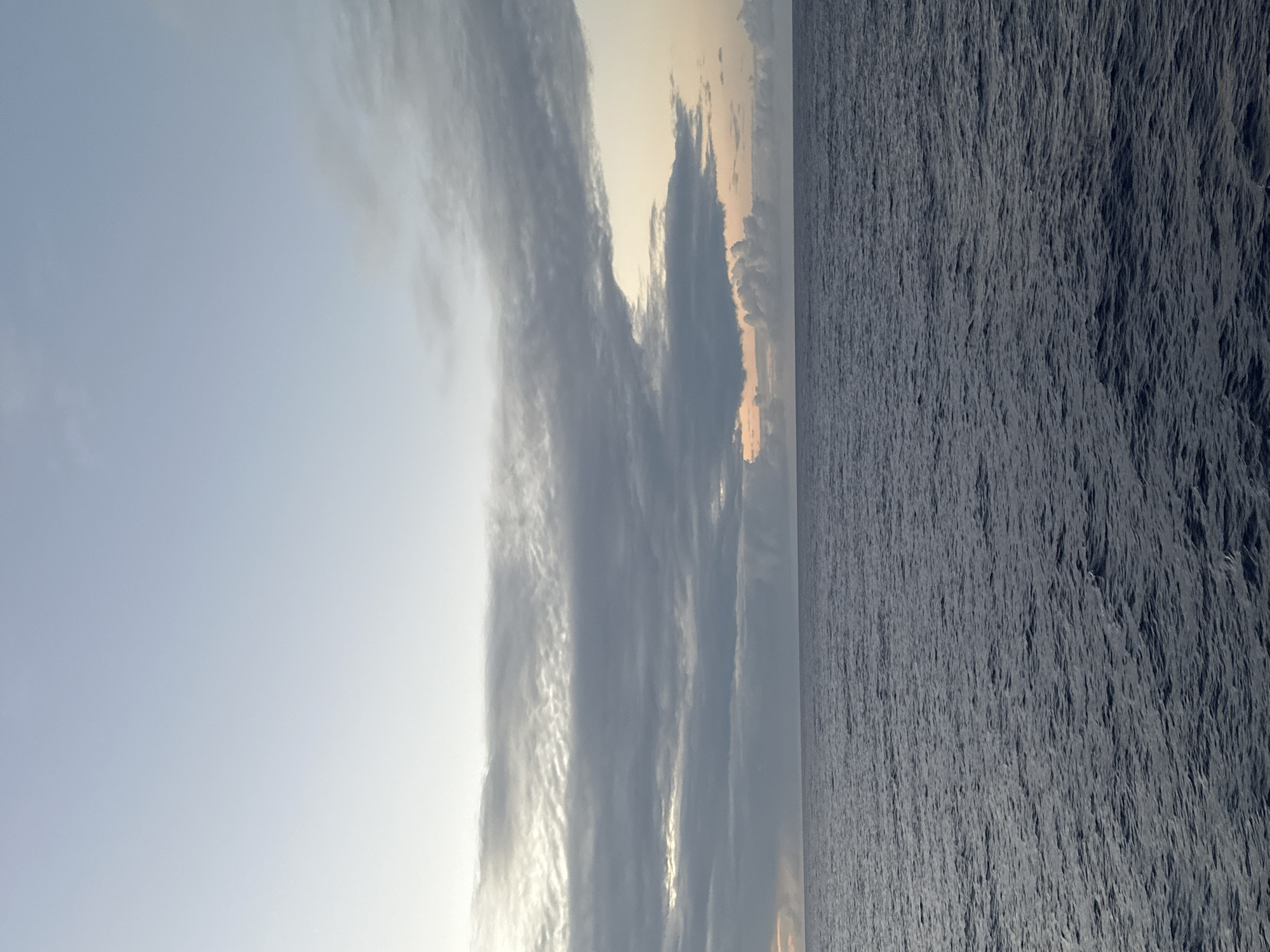 The image depicts a seascape with the sky and sea divided roughly in half. The sky is a pale blue with streaks of grey and white clouds that appear vertically oriented. Towards the upper right, the sky fades into a softer, warmer tone, possibly indicating sunrise or sunset. The sea occupies the right side of the image and is a uniform, dark blue-grey color. The surface of the water is textured with small ripples and waves, suggesting a slight breeze. The location is Unknown, Unknown.