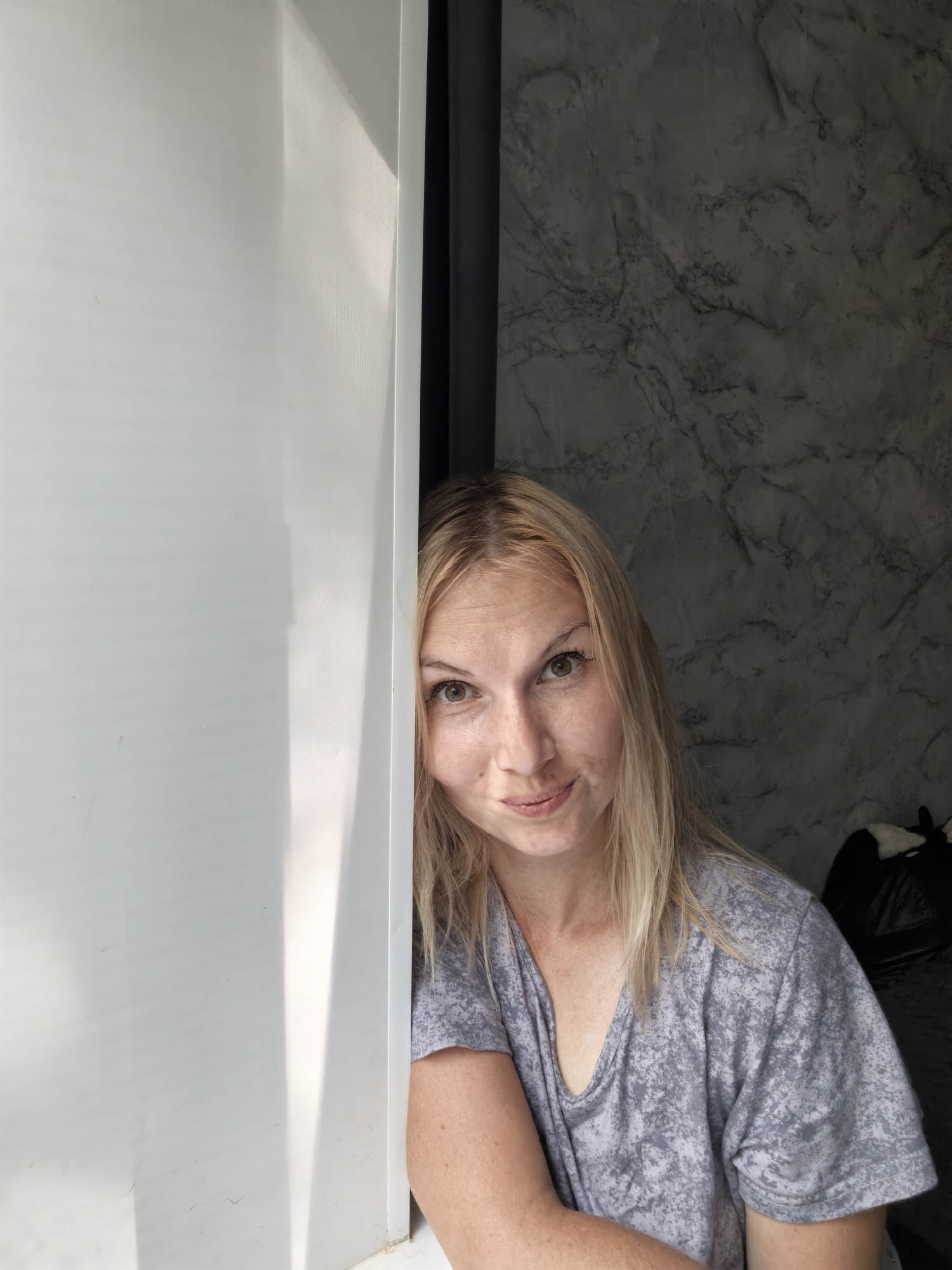 This is a portrait of a woman with blonde hair and light skin. She is looking at the camera with a slight smile and her head is tilted. Her eyes are green. She is wearing a grey t-shirt with a textured pattern. The woman is partially obscured by a white object on the left side of the frame, which appears to be a wall or door. Behind her, the background is a dark grey wall with a marble-like pattern. Sunlight is casting a shadow on the white object. The overall lighting suggests it is daytime. The woman's expression is relaxed and approachable. The scene appears to be indoors, possibly in a home or a casual setting. No text is visible in the image.