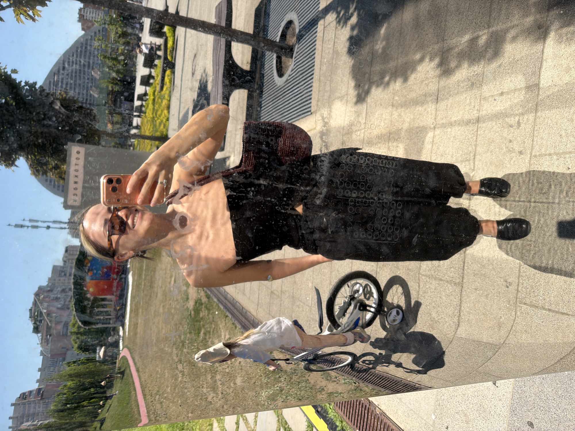 A woman is depicted taking a selfie, reflected in a curved, highly polished, vertical metallic surface under clear blue skies in Khimki, Russia. She smiles, wearing amber-framed sunglasses, a black strapless top, textured black wide-leg pants, and black flat shoes. She holds a rose gold smartphone in her right hand, capturing her own reflection, with a dark red woven bag slung over her left shoulder and her left hand in her pocket. The reflective surface is visibly smudged with circular marks, particularly across her upper body. To her left, also reflected, a young child wearing a light-colored hat and shirt is seen from behind, riding a white bicycle equipped with training wheels on a paved pathway.
The reflected environment behind them includes a grassy park area with a winding red path, scattered trees, and distant multi-story residential buildings on a hillside to the left. To the right, behind the woman, the reflection shows a paved plaza with dark benches, additional trees, and a large, distinctive wave-shaped modern building (consistent with structures like Arena Khimki) in the background. A reversed Cyrillic sign, appearing to read "ФОТОБОКС" (Photobox), is visible in the upper right of the reflection. The ground immediately in front of the reflective surface is paved with light-colored tiles, and prominent shadows indicate strong overhead sunlight.