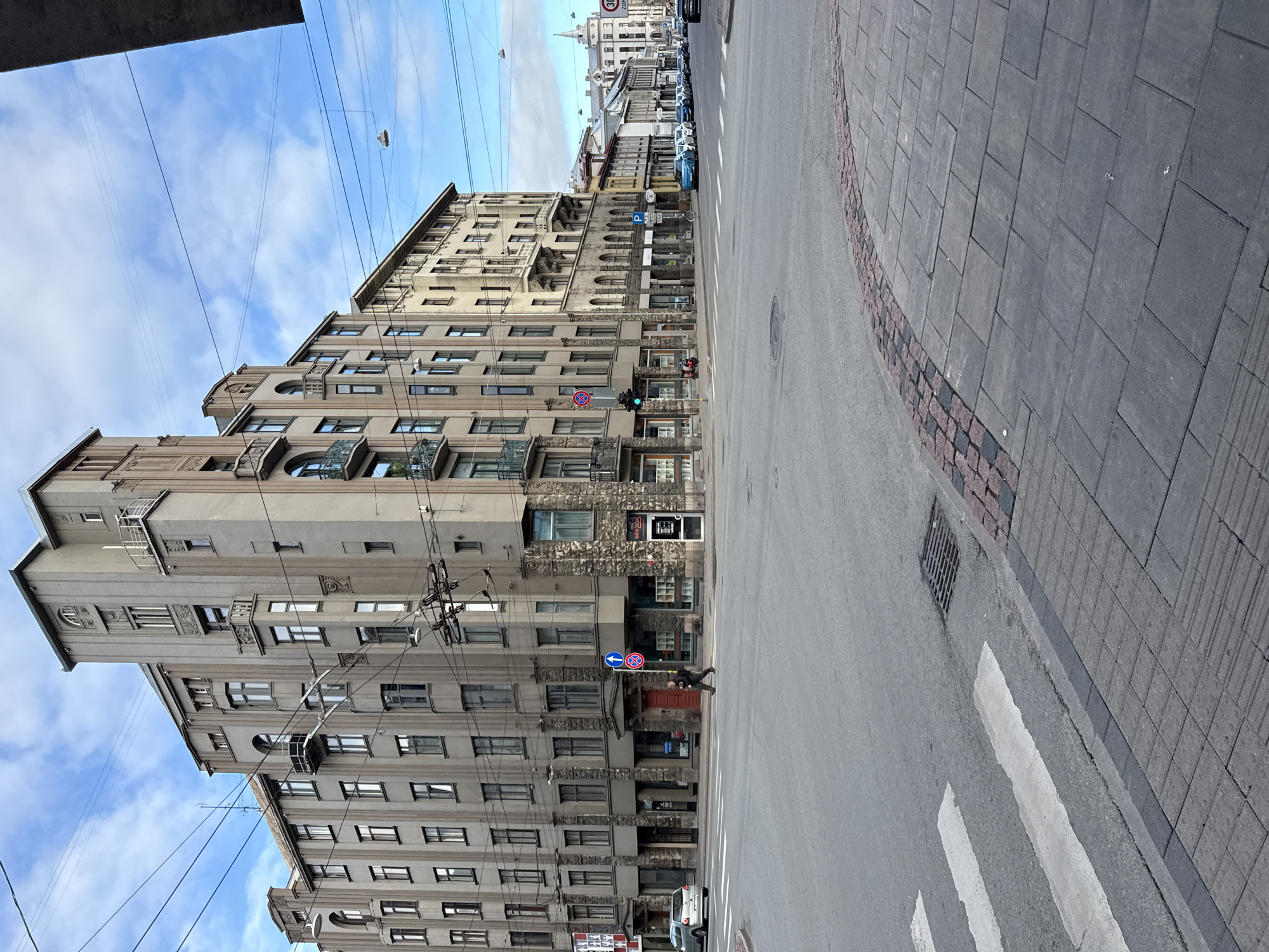 The image shows a street intersection in Riga, Latvia. The main focus is a large, multi-story building on the corner, characterized by its gray facade, numerous windows, and decorative architectural elements, including balconies and stone-clad lower levels. Several other buildings line the street, appearing older and with lighter-colored facades. A pedestrian, dressed in dark clothing, is crossing the street at a marked crosswalk. Other vehicles, including a silver car and several parked cars further down the street, are visible. Overhead power lines crisscross the sky, typical of urban European cities. The sky is partly cloudy with a blue sky visible. The street is paved with asphalt, and the sidewalks are made of stone pavers. A traffic light is visible and appears to be red for the direction of the pedestrian. A street drain is present on the side of the road. The image captures a typical city scene with a blend of old and new architecture and urban infrastructure.