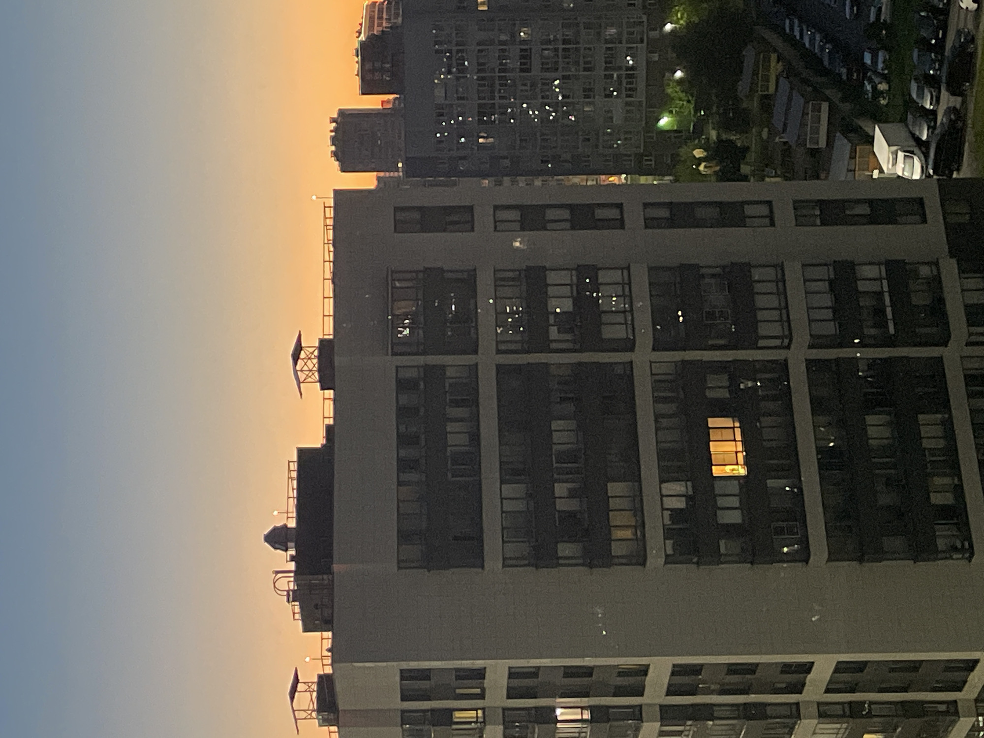 A twilight scene captures a cluster of tall residential buildings in the city of Sinyavino, Russia. The sky transitions from a pale blue at the top to a warm orange hue near the horizon, indicating sunset or sunrise. In the foreground, a multi-story apartment building with a light-colored facade and dark-paneled sections dominates the view. Numerous windows, some illuminated with warm yellow light, punctuate the building's structure.
Behind the foreground building, other apartment complexes rise, their facades darker and also dotted with lit windows, creating a sparkling effect. To the right, a row of parked cars is visible, including a white van. The ground level reveals patches of greenery and what appear to be small garden structures or sheds. The overall impression is of a densely populated urban environment during the evening or early morning hours.