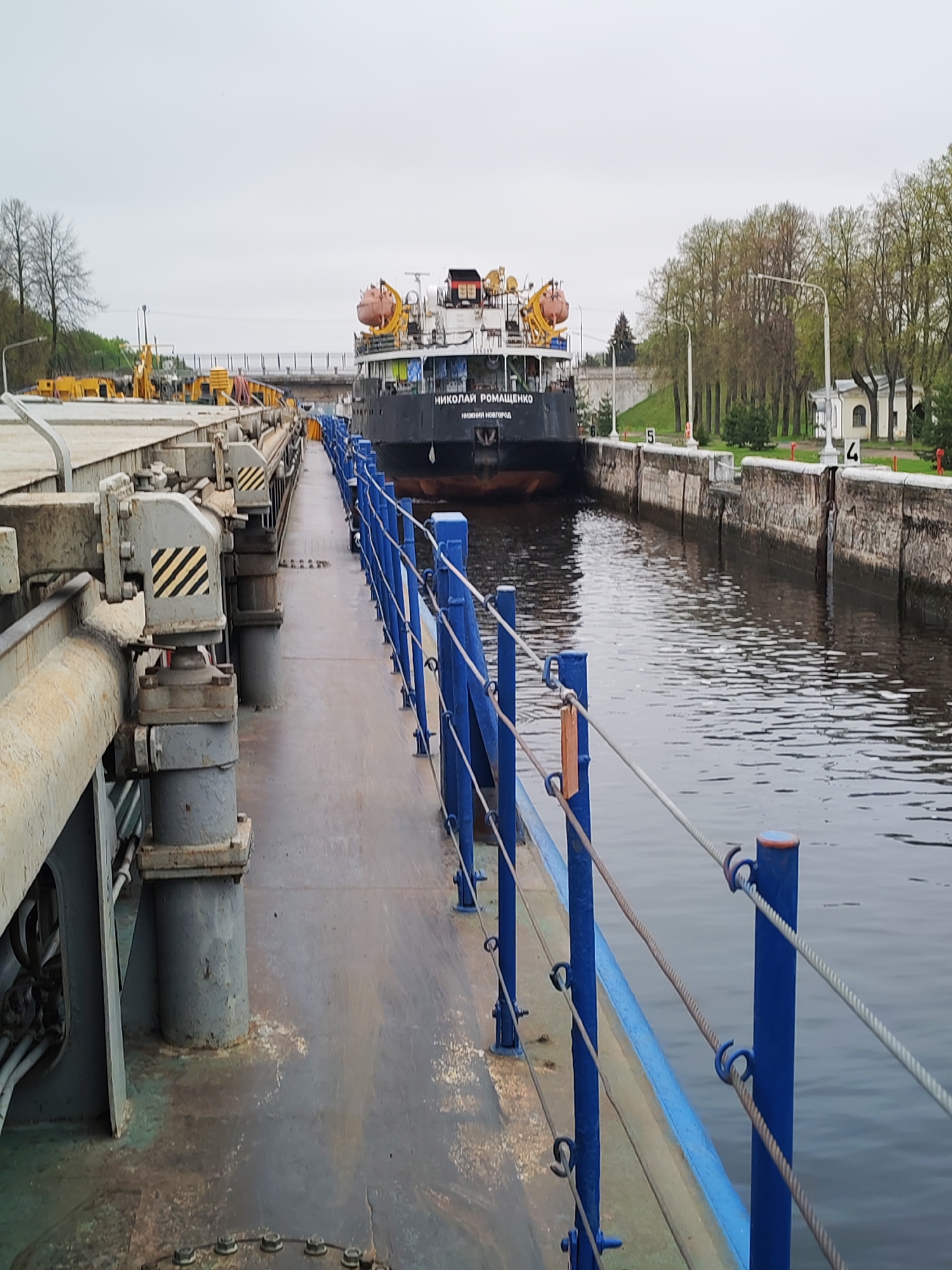 The image depicts a canal with a large vessel named "Николай Ромащенко" situated within a lock. The lock walls are numbered "4", "5", and "6". A walkway extends along the side of the canal, featuring blue railings with ropes. Heavy machinery with black and yellow hazard stripes is visible on the left side of the walkway. Trees and a building are visible in the background on the right. The location is a lock on a canal, and the sky is overcast. The city is not apparent from the image.