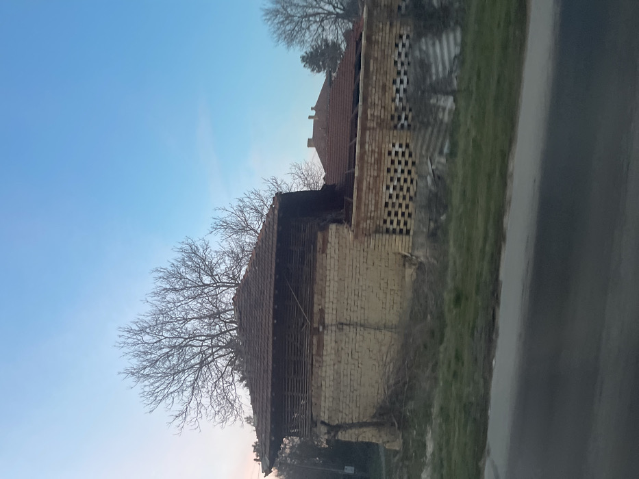 The image depicts a rural scene in Osenets, Bulgaria, featuring a dilapidated two-story structure under a clear sky. The primary building has a lower level constructed of light-colored bricks, showing signs of decay and crumbling sections. Its upper level consists of horizontal wooden slats under a gabled roof made of red clay tiles, which exhibits partial collapse and missing sections on the right side. Behind this structure, the skeletal branches of bare deciduous trees are visible. To the right, an attached or adjacent wall of red bricks features an open lattice pattern, with a partially visible tiled roof behind it, indicating another structure or extension in disrepair. The immediate foreground shows a dark asphalt road bordered by a strip of green grass and dry brush. Scattered debris, including corrugated sheets, lies within the grassy area. No individuals are present. The sky is a gradient of light blue transitioning to a faint pinkish hue near the horizon.