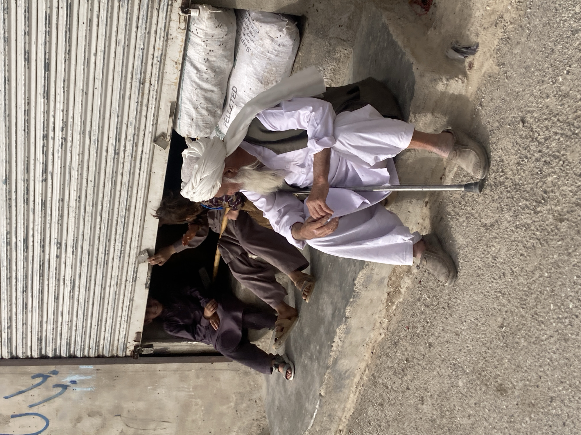A low-angle shot shows an elderly man with a white beard, wearing a white turban and traditional white clothing, using a metal walking stick to support himself. He is sitting on the ground next to a closed metal shutter door. Behind him are two large white sacks labeled "PELLETED." Two younger individuals are visible emerging from under the shutter door. One is wearing brown clothing, and the other is wearing black clothing. The scene is located outdoors on a paved surface in Quetta City Tehsil, Pakistan.