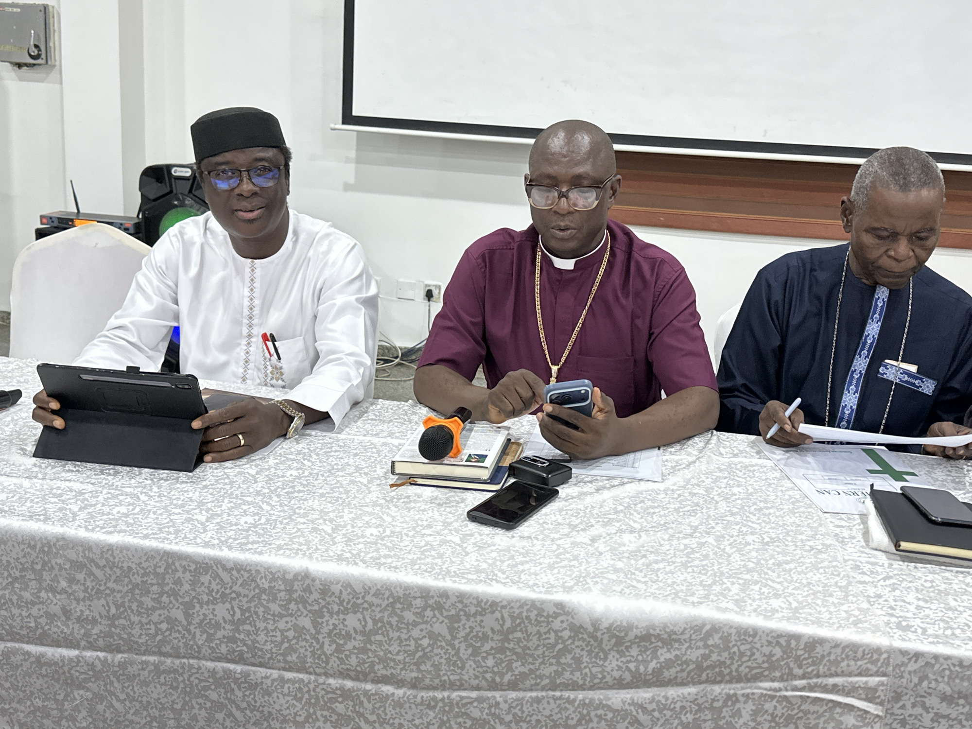 The image depicts three individuals seated at a table covered with a white and grey patterned cloth. The setting appears to be a conference room or meeting space, possibly within a religious or governmental institution in Abuja, Nigeria.
The person on the left, wearing a black cap and a white traditional attire, is looking towards a tablet in front of them. They have glasses and are wearing a watch. In front of this person are pens in a pocket.
The central individual, who appears to be a clergyman, is wearing a dark burgundy clerical shirt with a white clerical collar and a gold chain. They are looking down at their mobile phone, which they are holding in their right hand. In front of them are stacked books, a microphone, and another mobile phone.
The person on the right, also appearing to be a clergyman, is wearing a dark blue traditional attire with intricate white and blue embroidery on the collar and chest. They are looking down at papers in front of them and holding a pen in their right hand. There are documents and notebooks on the table in front of this individual.
The background shows a white projector screen, electrical outlets, and some audio equipment. The overall lighting suggests an indoor environment. The table is set with various items related to a meeting or presentation.