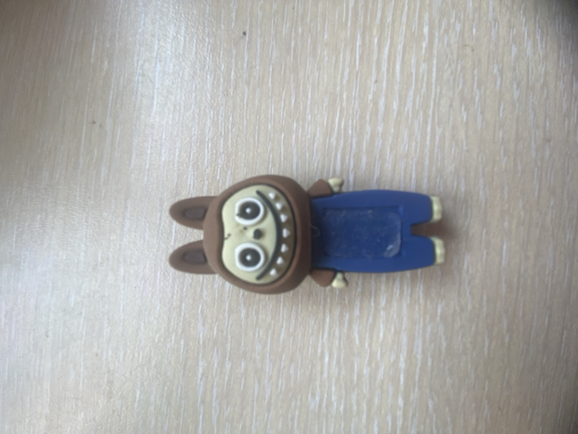 A small, brown, cartoonish figurine is centered on a light-colored, wood-grained surface. The figurine has large, white-ringed eyes with black pupils, and a wide, toothy smile with sharp, triangular teeth. It wears a blue jumpsuit and has brown paws or hands. The figurine is standing upright. The surface appears to be a table or desk. The image does not provide any information about the city of Blagoevgrad, Bulgaria.