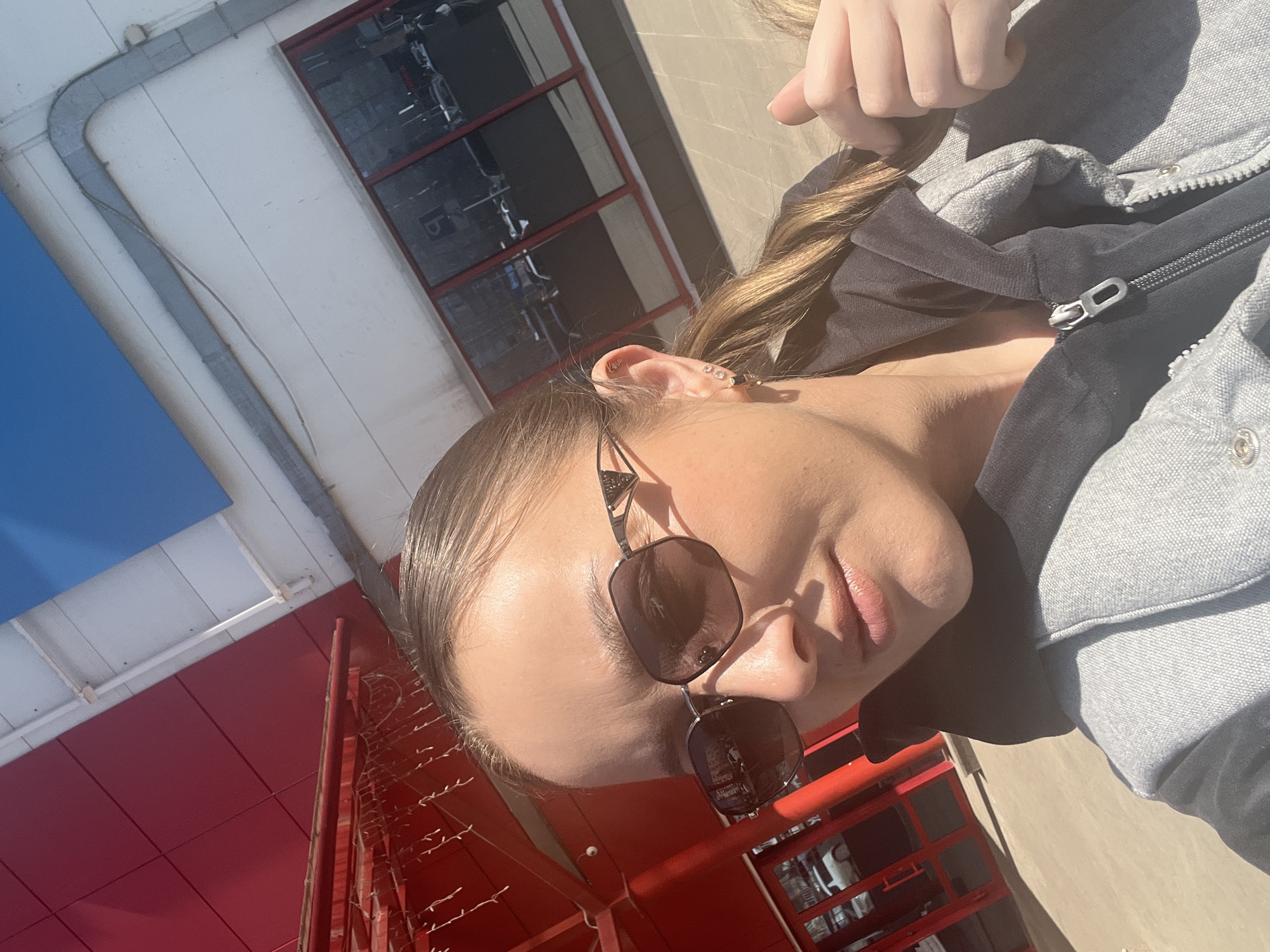 A woman wearing sunglasses and a gray jacket is posing outside a building. The building appears to have a red and white exterior, with windowed sections showing what seems to be a fitness center inside. The photo is taken at an angle, making the scene appear slightly tilted. The location could be Izhevsk, Russia, based on the information given.